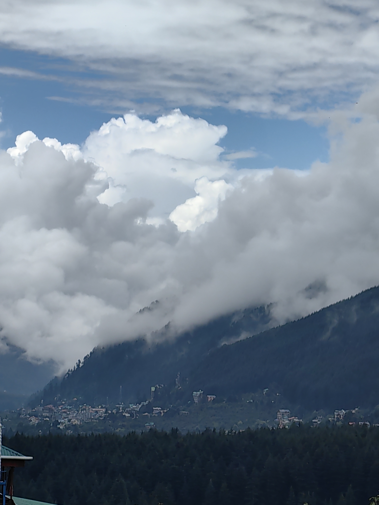 The image depicts a panoramic view of a mountainous landscape, likely Vashisht, India, characterized by a dense forest in the foreground and a town nestled on a hillside in the background. The sky is filled with dramatic clouds, with patches of blue visible, suggesting a dynamic weather system. The town appears to be composed of multi-story buildings with varied architectural styles, clustered together on the slope of the mountain. A significant portion of the mountainside is covered by a dark green forest, with a distinct line of trees demarcating its edge. Some parts of the mountain are obscured by the clouds, creating a sense of depth and atmosphere. In the very bottom left corner, a portion of a structure with a green roof is visible, suggesting a vantage point from which the photograph was taken. There are no people or animals visible in the frame.
