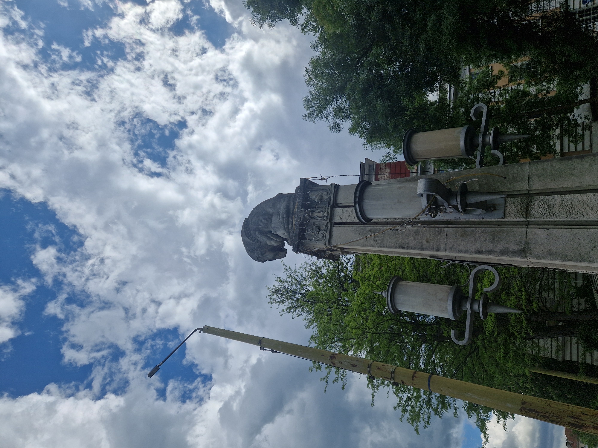 The image shows a monument topped with a bust of a person, possibly made of stone, situated in an urban environment. The sky is visible, filled with white and gray clouds. Trees are present, their foliage appearing green. The monument has ornate designs and is fitted with lamp structures designed to look like syringes. A tall, yellow pole with a light fixture at its top extends upward, parallel to the monument. The location is in the city of Unknown, Unknown.
