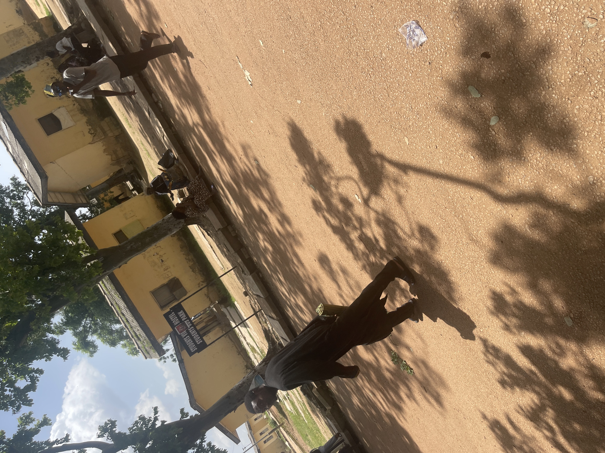 The image depicts an outdoor scene, likely on a street or pathway, with a sandy or dirt ground. In the foreground, a man wearing dark clothing and sandals is walking towards the left of the frame. He is carrying a cylindrical object in his right hand. In the mid-ground, another man in a grey t-shirt and dark pants is running or walking quickly to the right, wearing sunglasses and a yellow cap. Several other people are visible in the background, some sitting on a bench and others standing near yellow buildings. A signpost reads "STUDENTS UNI HOSTEL STUDENTS BUS PARK". The environment appears to be somewhat weathered, with some buildings showing signs of age. Shadows of trees are cast across the ground, indicating a sunny day. There is some litter, including a plastic bag, visible on the ground. The location is identified as Rafin Par, Nigeria.