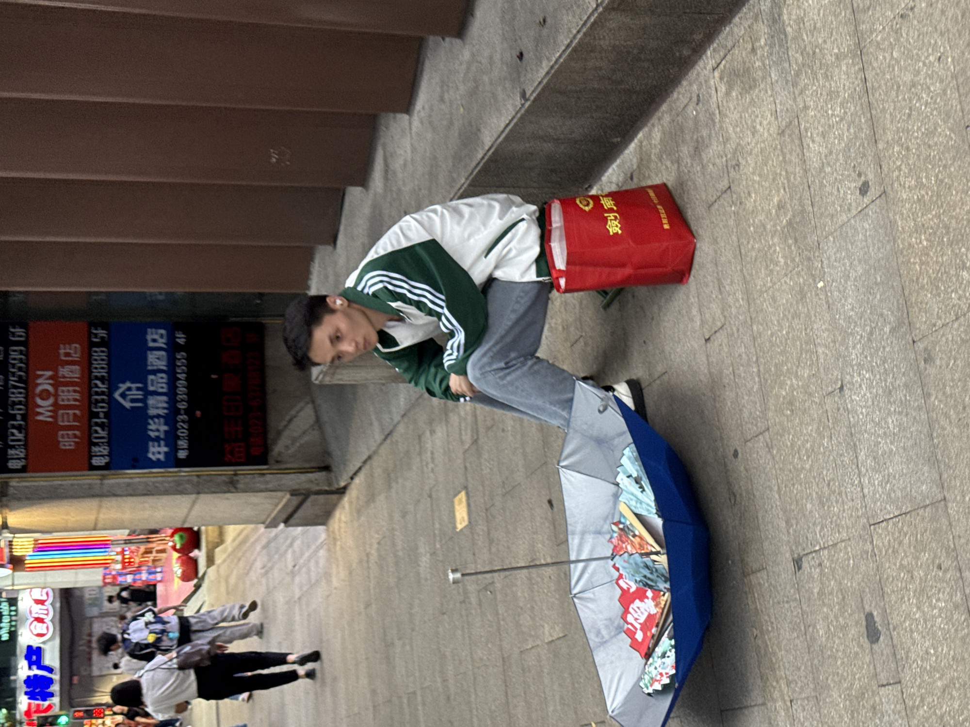 A person in a green and white jacket is squatting next to a red bag on a street in Yuzhong District, China. A blue umbrella lies partially open on the ground. Behind the person are signs with Chinese writing and telephone numbers. People are walking in the background. The signage advertises hotels.