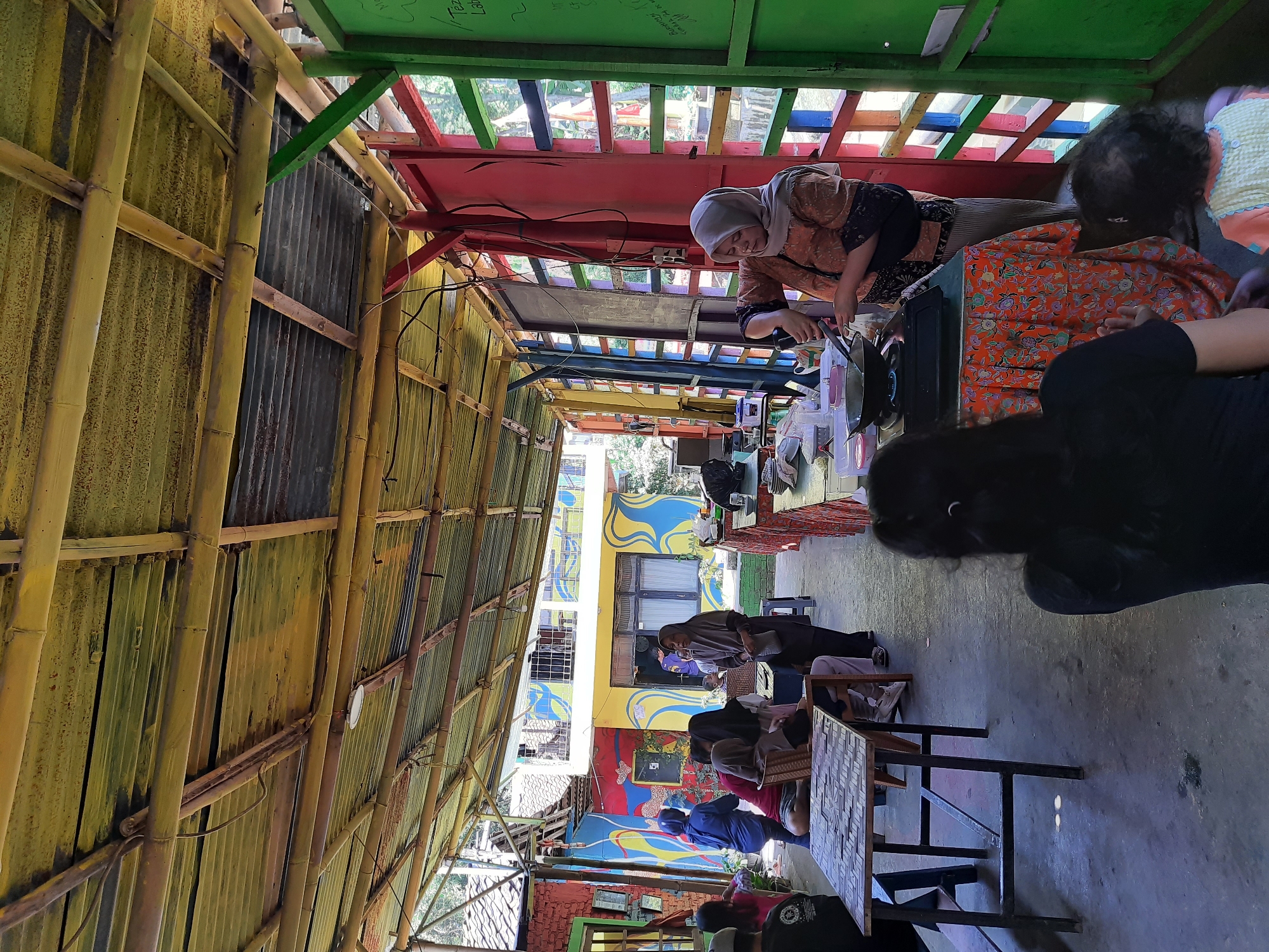 The image shows an outdoor cooking demonstration at a colorful, open-air venue. A woman wearing a hijab and an orange patterned top is actively cooking on a portable stove, stirring a pan with a spatula. She appears to be demonstrating a recipe to others. Several people are seated at tables, observing the demonstration. The environment is vibrant, with a bamboo-and-corrugated metal roof overhead, painted walls with bold abstract designs, and multi-colored window frames. In the background, a building with similar colorful murals is visible. The ground is a concrete floor. The scene suggests a community event or workshop.