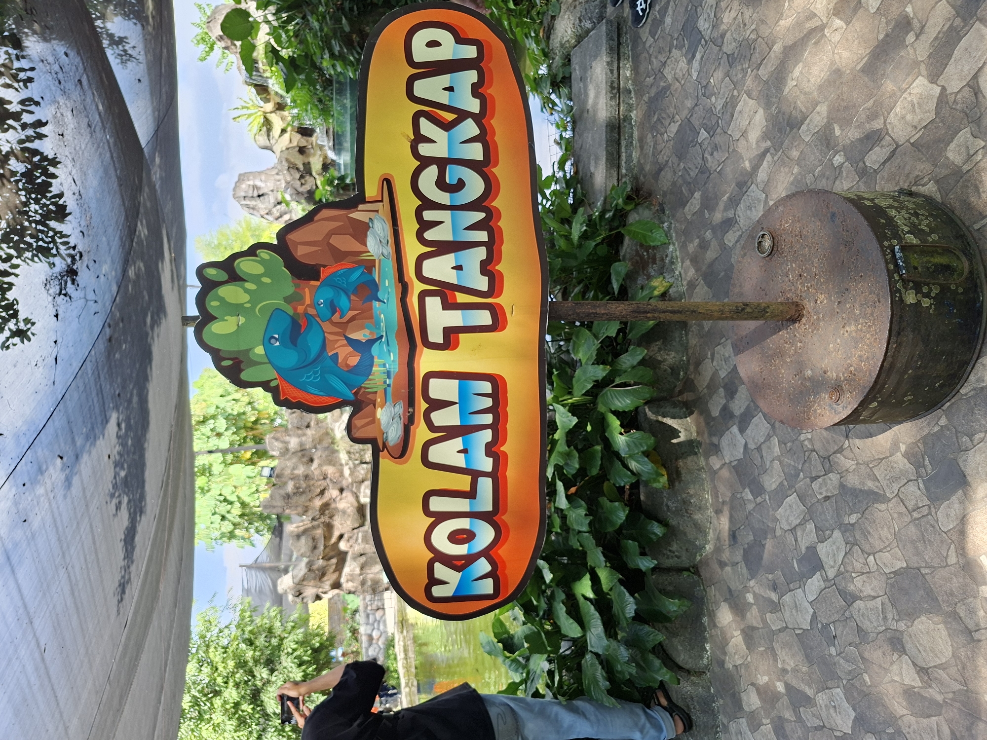 The image captures a sign that reads "KOLAM TANGKAP" in large, stylized letters, indicating a fishing pond or catch pool. The sign is yellow with orange trim and features cartoon depictions of two blue fish. It is mounted on a rusty metal pole, which is secured to a weathered metal base. The setting appears to be an outdoor attraction, possibly a zoo or a family entertainment center, given the presence of lush greenery, artificial rock formations, and a body of water in the background. The ground is paved with irregular stone tiles. A person wearing a black shirt and blue jeans is visible to the left, holding a smartphone and seemingly taking a photo. The overall impression is that of a casual, recreational environment, likely in a warm climate given the presence of broad-leaved plants and a covered, open-air structure above. The sign's language suggests the location is in Indonesia.