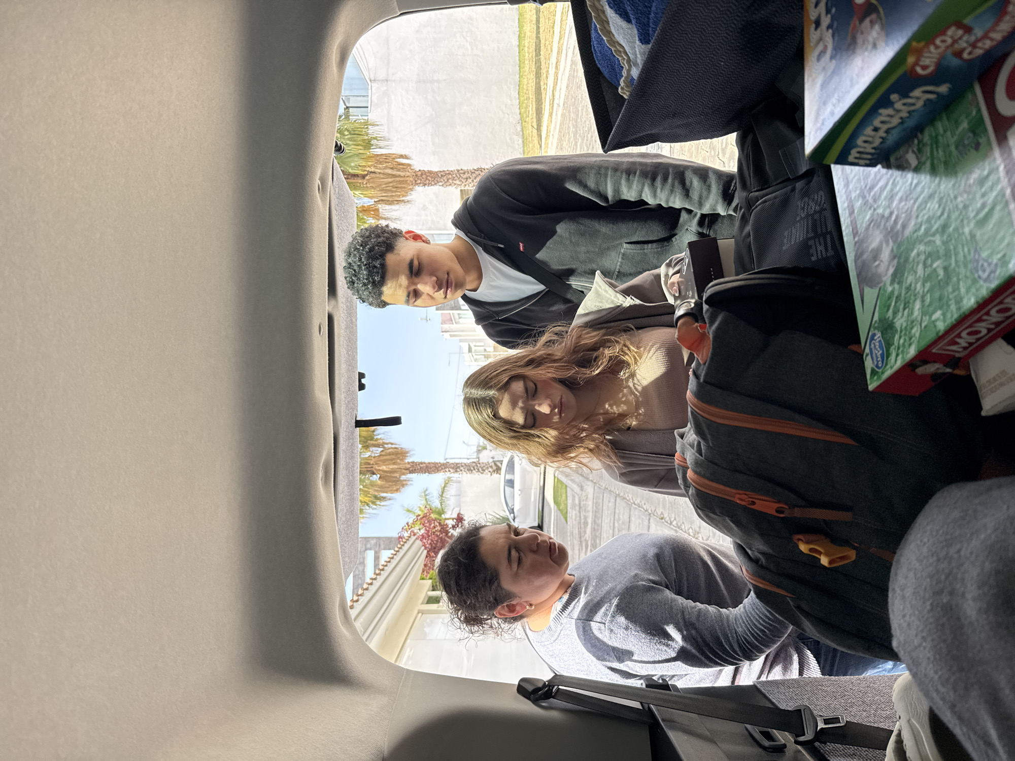 The image shows three people in the open trunk of a vehicle. A young man stands at the front of the trunk looking down, wearing a black hoodie over a white t-shirt. Behind him, a young woman with long blonde hair looks down as well, wearing a neutral-colored sweater. At the rear of the trunk, a person with short curly hair is seated, wearing a gray long-sleeved shirt. The trunk contains luggage including a backpack and several board game boxes, including Monopoly and Marathon. The background shows a sunny outdoor scene with palm trees and buildings, suggesting the location is San Bernardino Tlaxcalancingo, Mexico.