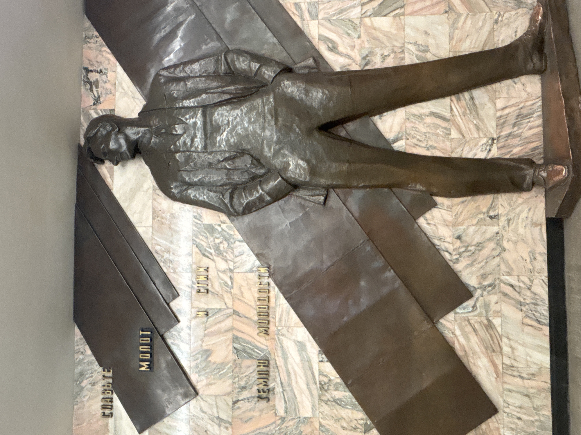 The image is a bronze sculpture of a man lying on a wall with a marble pattern. The sculpture is located in Saint Petersburg, Russia. The man is wearing a suit and is positioned diagonally across the wall. The sculpture is part of a monument titled "Glory to Labor," located in the Kirovsky district. There are bronze rectangles at the bottom left of the sculpture, with words inscribed on them. The words are in Cyrillic alphabet.