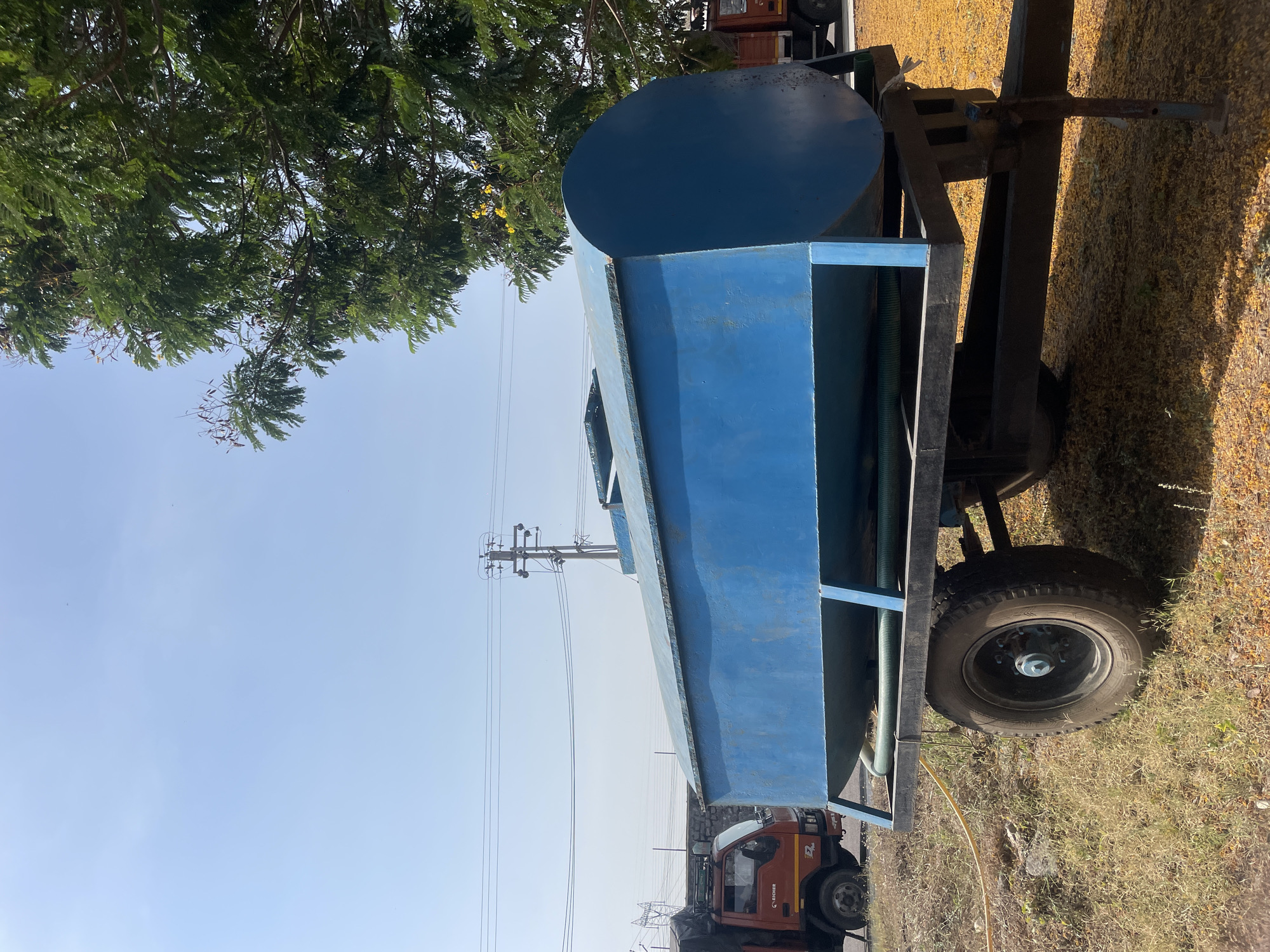 The image depicts a blue water tank mounted on a trailer with wheels, situated outdoors in what appears to be a rural or semi-urban area. The tank is cylindrical at one end and rectangular along its length. The trailer's frame is black. The trailer is parked on dry, grassy ground. In the background, power lines and a utility pole are visible. Also, a red truck is partially visible. The presence of the truck and utility pole suggests a location within or near a populated area. The city of Anajpur, India is presumed to be the location based on the prompt instructions. There are some green leaves on the top left corner.