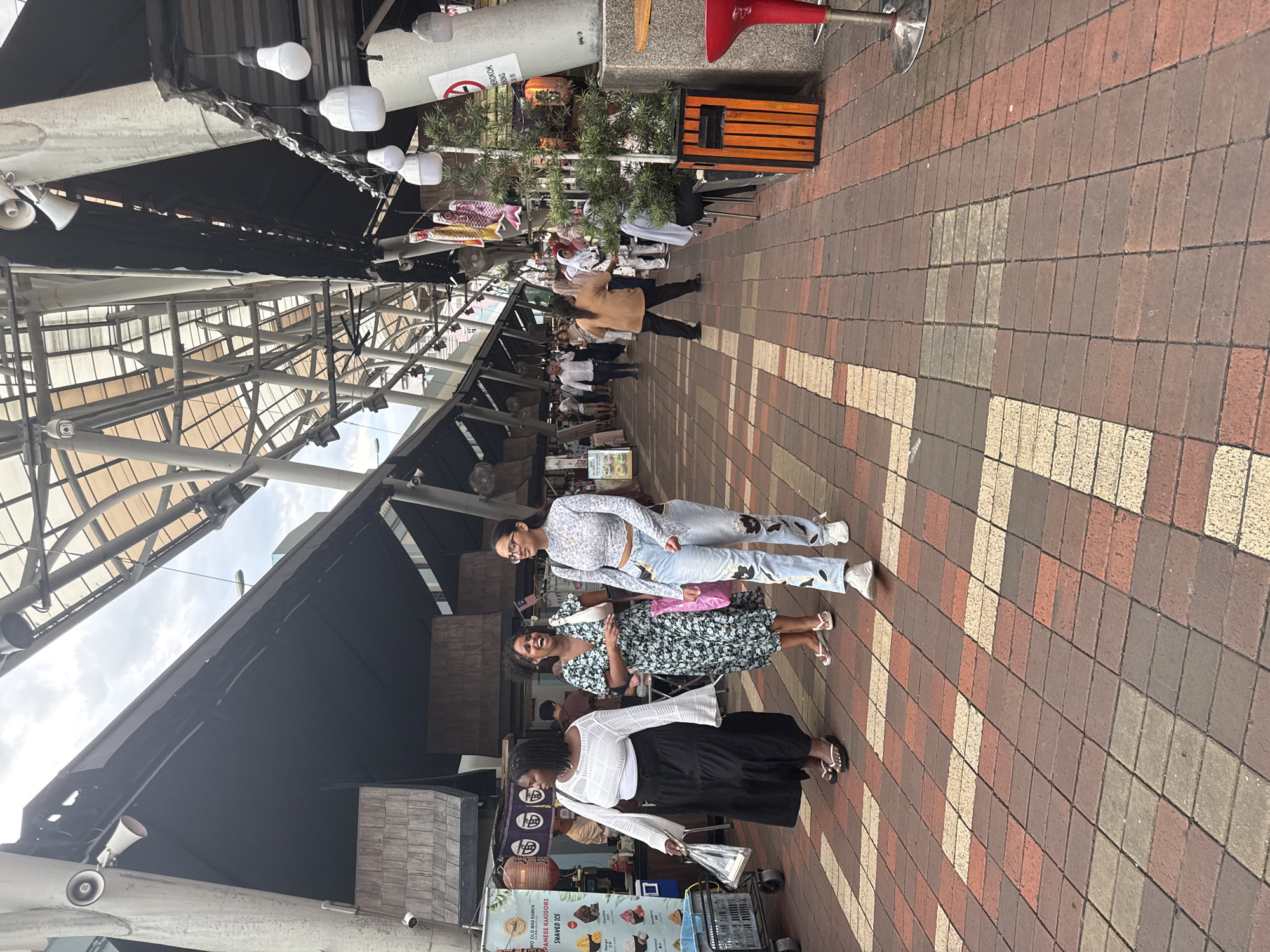 Three women stand on a brick walkway under a metal and black canopy, likely in Kuala Lumpur, Malaysia, given the presence of Malaysian flags. One woman wears a white top and a long black skirt and holds a clear plastic bag. The second woman wears a floral dress, while the third is in a floral top and light-wash jeans. A food stall featuring "Japanese Old Man Ramen" is visible near the women. Other pedestrians are walking in the background. Decorations, including rows of lightbulbs and wind socks, are strung above.