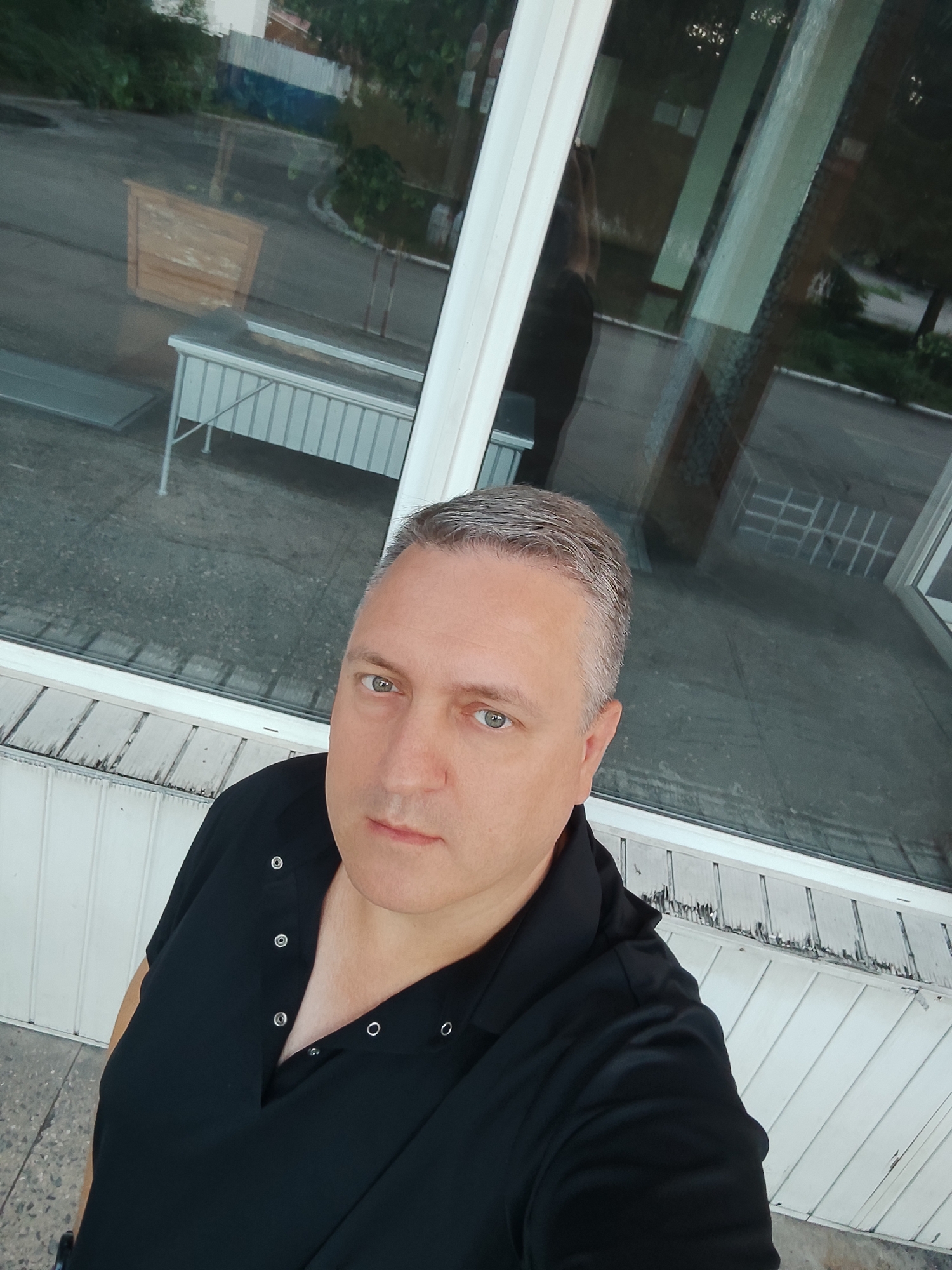 An outdoor, waist-up, selfie shows a light-skinned, middle-aged man with graying hair, wearing a black collared shirt with snap buttons, standing outside in front of a large window. The man's head is tilted slightly to the right, and he is looking directly at the camera with his blue-green eyes. The window reflects an outdoor scene with a road, trees, and buildings. The man is positioned in the foreground, with the window and its reflections taking up the background. The lower part of the image shows a white wooden structure. The lighting suggests it is daytime, with overcast or diffused light.