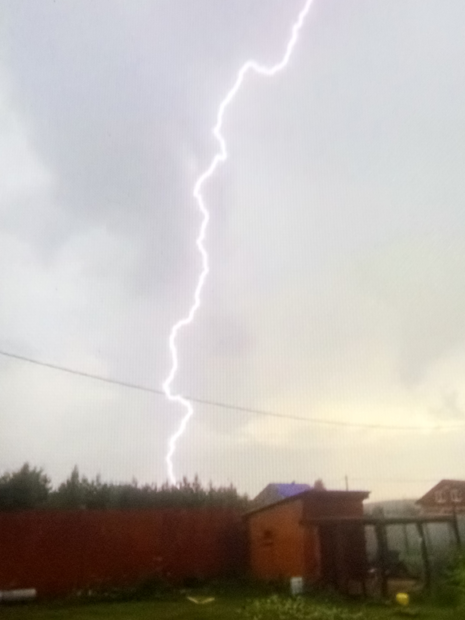 The image depicts a dramatic sky filled with a bright lightning strike during a thunderstorm. In the foreground, a dark red fence runs horizontally across the frame, with a patch of green grass and vegetation visible below it. Behind the fence, a cluster of trees forms a dense line. On the right side of the image, there are several buildings, including what appears to be a small brick structure with a dark roof and a larger building with a peaked roof. The sky is predominantly gray and overcast, with the exception of the intense white light of the lightning bolt that zigzags from the upper portion of the image down towards the horizon. The overall impression is one of a powerful natural event occurring over a rural or suburban landscape.