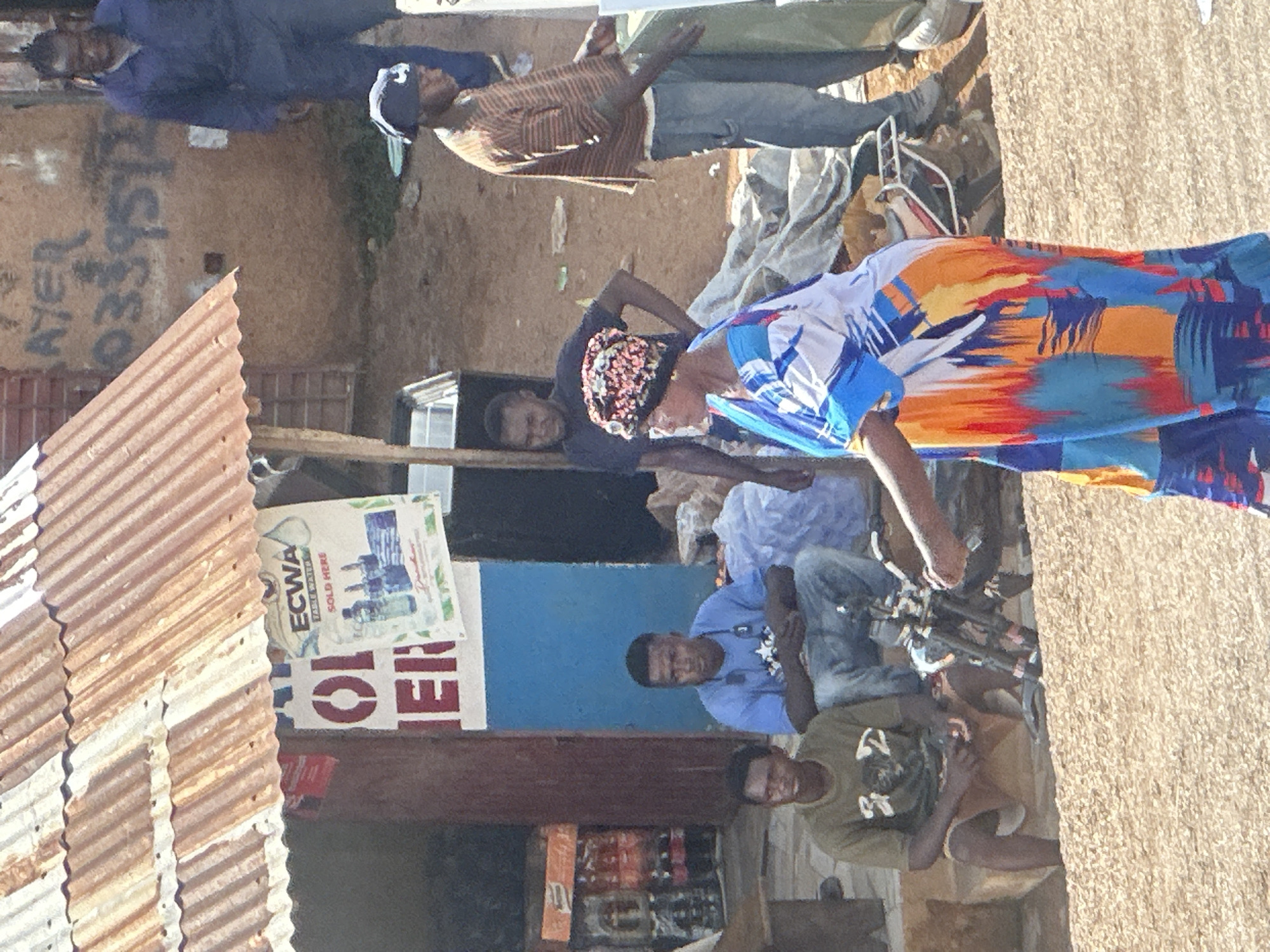The image depicts a street scene in Jos, Nigeria, likely a commercial area with an open-air market or shopfront. A corrugated iron roof, aged and rusted, partially shelters the front of a building. Two men are seated in front of the building. The man on the left wears a greenish-brown t-shirt and brown shorts, while the man on the right is in a blue t-shirt and blue jeans.
A woman in the foreground is wearing a colorful, patterned dress and a dark headscarf. She is looking to the right. To her right, another man in a striped brown and orange shirt and blue jeans is standing and gesturing with his right hand towards something out of frame to the right. Behind him, a man in a dark t-shirt is leaning against the wall. Further back and to the left, a man in a blue work uniform is walking away from the camera.
Various objects are visible: a motorcycle is parked on the ground in the middle ground, and a stack of plastic crates filled with bottles are visible to the left of the building. A poster advertising "ECWA TABLE WATER SOLD HERE" is attached to the front of the building. The ground is a dusty, uneven surface, consistent with unpaved areas in many Nigerian cities. Graffiti is visible on the wall behind the standing man in the striped shirt.
