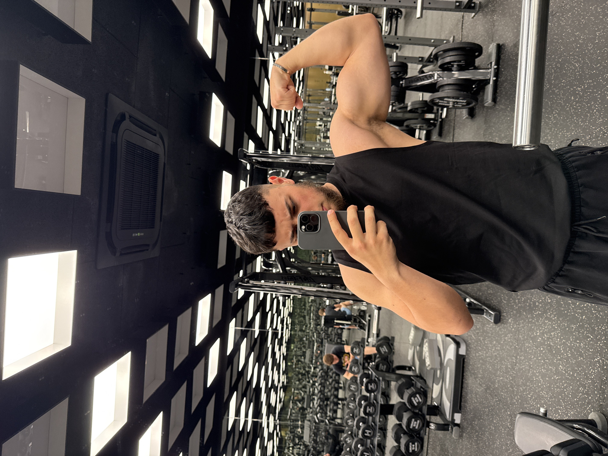 The image is a self-portrait of a man flexing his bicep in a gym. He is wearing a black tank top and black shorts. The gym appears to be well-equipped with various weight machines and dumbbells, and the lighting suggests it is an indoor space. The ceiling has a modern design with recessed lighting panels and what looks like an air conditioning unit. The floor is a speckled rubber mat, typical of gyms. In the background, other gym-goers can be seen exercising. The overall scene depicts a typical gym environment focused on fitness and weight training. The location is not specified in the image or its metadata, therefore it cannot be stated that this image was taken in Varna, Bulgaria.