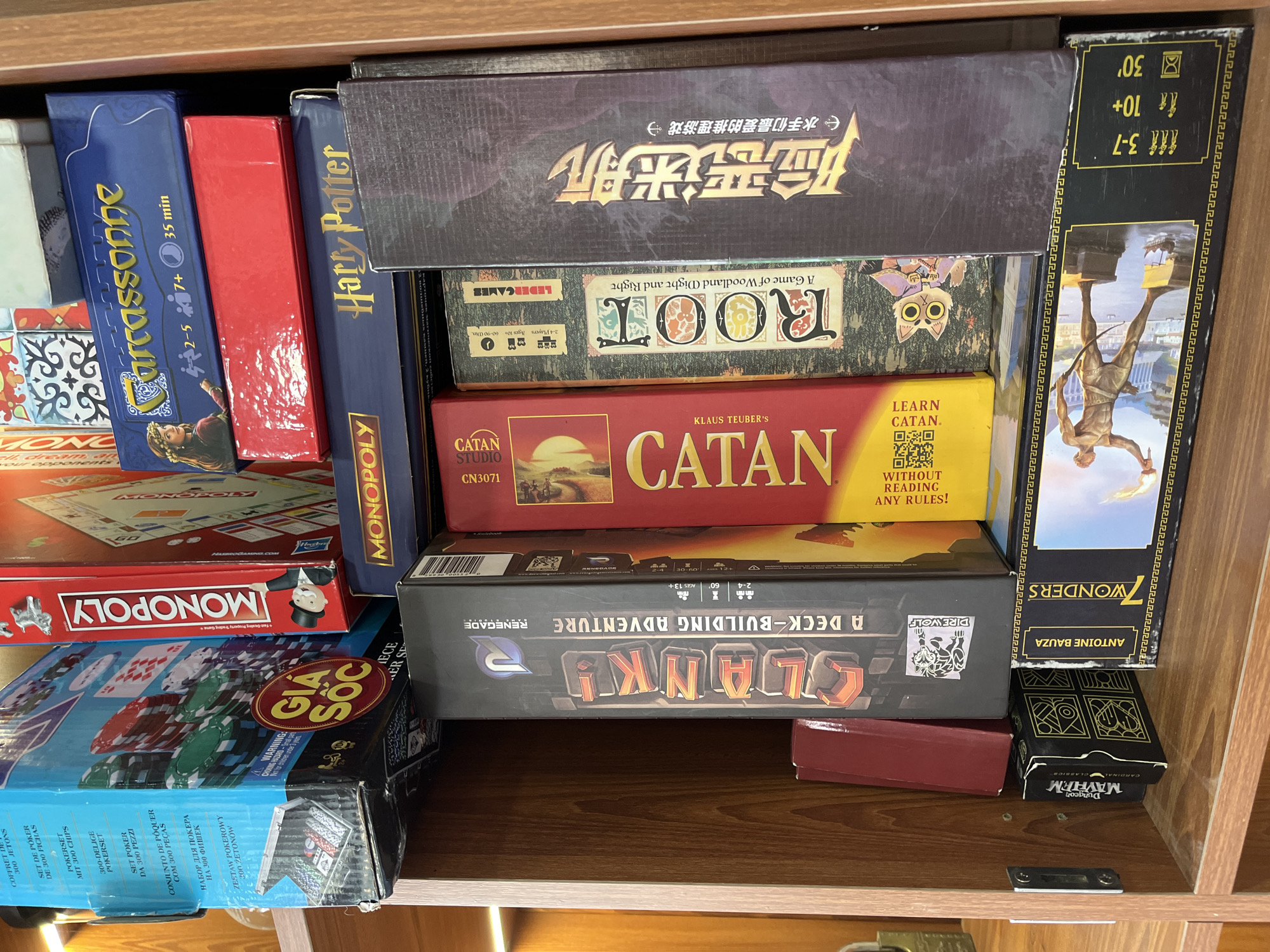 The image shows a collection of board games stacked on a shelf inside a wooden cabinet. The games include: "Harry Potter," "Carcassonne," "Monopoly," "Gia Soc," "Slank!," "7 Wonders," "Catan," "Root," and "Dungeon Mayhem." The shelf appears to be in a residential setting. There is no visual information to determine the location in Đà Nẵng, Vietnam.