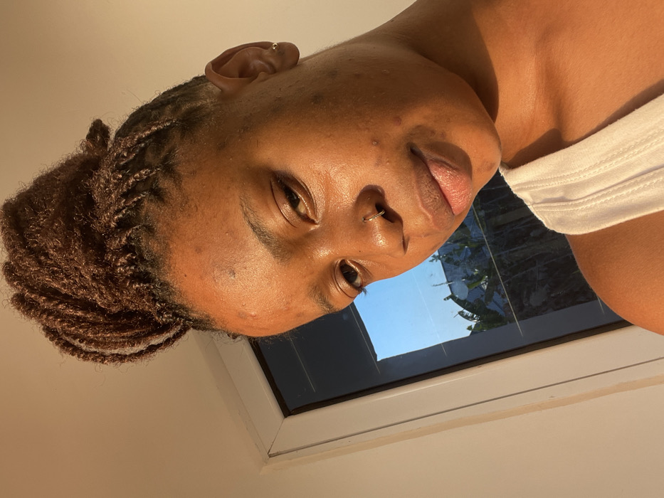 A person poses for a selfie indoors, illuminated by warm light. They have dark skin, brown hair styled in small twists piled atop their head, and facial piercings in their nose and ear. A white tank top is visible on their right shoulder. Behind them is a window with a partial view of blue sky and green foliage.