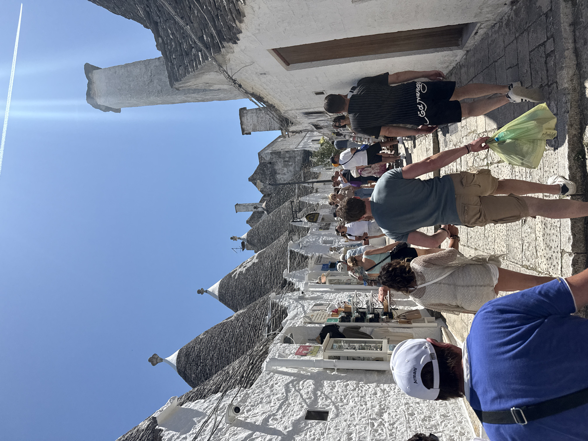 The image depicts a street scene in Alberobello, Italy, characterized by its distinctive trulli houses. These structures have conical roofs made of stacked limestone slabs. A group of tourists is present, walking along the cobblestone street, seemingly engaged in sightseeing and shopping. Some appear to be browsing at outdoor displays of merchandise, while others are simply passing through. The sky is clear and blue. An airplane contrail is visible in the upper left quadrant. The buildings are whitewashed with dark stone roofs. The perspective suggests the photographer is positioned amidst the crowd of tourists.