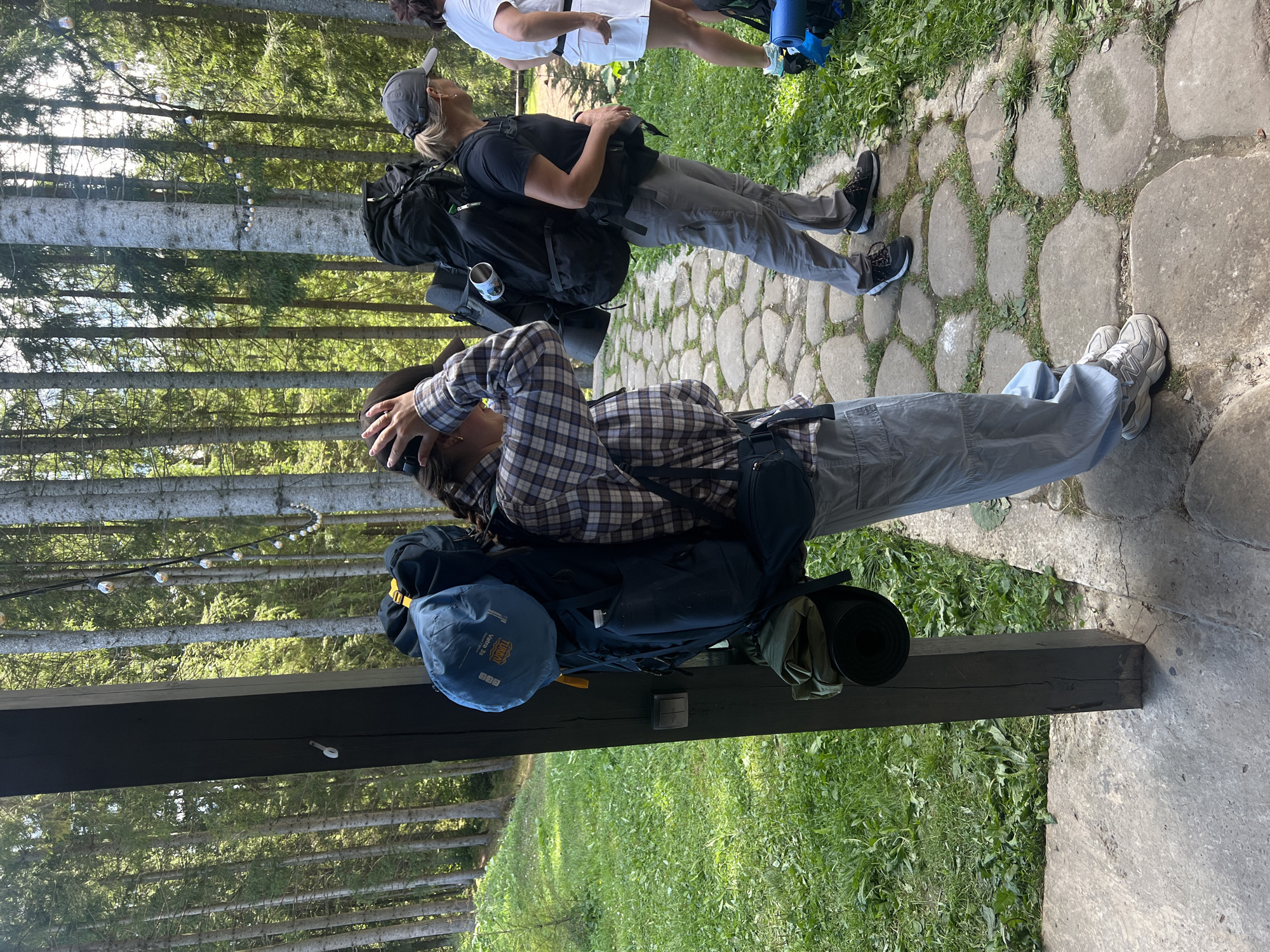 The image depicts a group of people with backpacks standing on a stone pathway in a forested area, likely in the city of Vorokhta, Ukraine. In the foreground, a person wearing a plaid shirt and light-colored pants is adjusting their hair while standing next to a large, dark blue backpack. Another person, wearing a dark t-shirt and gray pants, stands to the right, also with a large black backpack. A third person, wearing a white t-shirt and white shorts, is visible further to the right. The environment is characterized by tall trees, green grass, and a cobblestone path. Decorative string lights are visible strung between trees in the background, suggesting a recreational or camping setting.