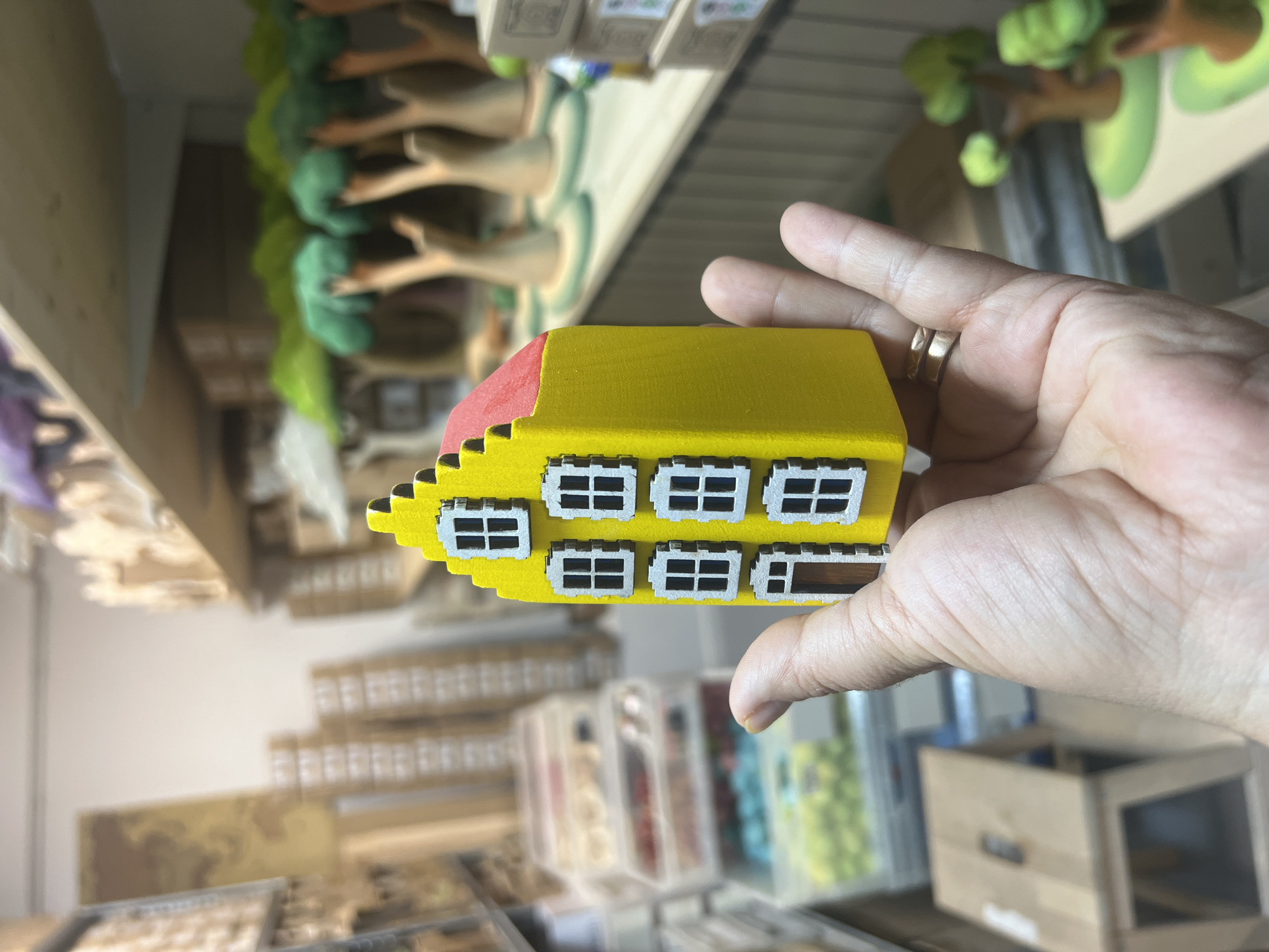 A person's hand holds a small, pixelated toy house. The house is yellow with a red roof and features white window frames and a brown door. The background is blurred but suggests a store or workshop setting, with shelves stocked with various items, including what appear to be more wooden toys, possibly trees, and boxes. There are also storage containers filled with colorful beads or craft supplies visible. The overall scene is brightly lit.