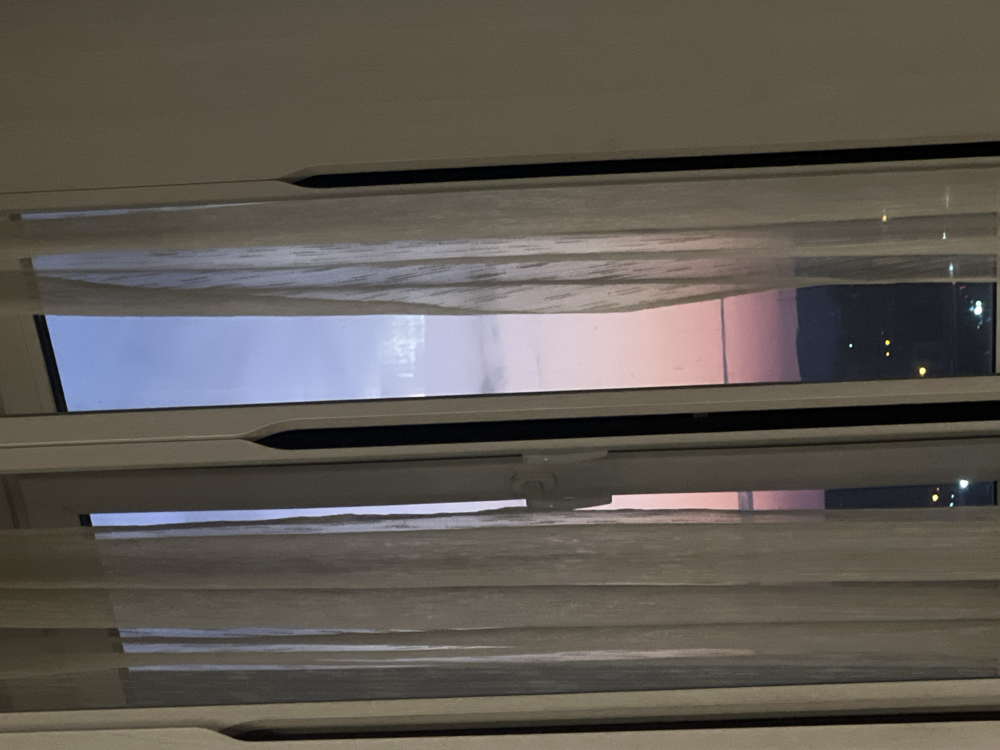 A view through a window with horizontal blinds, showing a transition from daylight to night. On the left, the sky appears light blue, gradually shifting to a pinkish hue towards the right, where it transitions to dark, with distant lights suggesting a cityscape at night. The window frame and blinds are light-colored. No people are visible. The location is unknown, but the prompt suggests Krasnoyarsk, Russia.