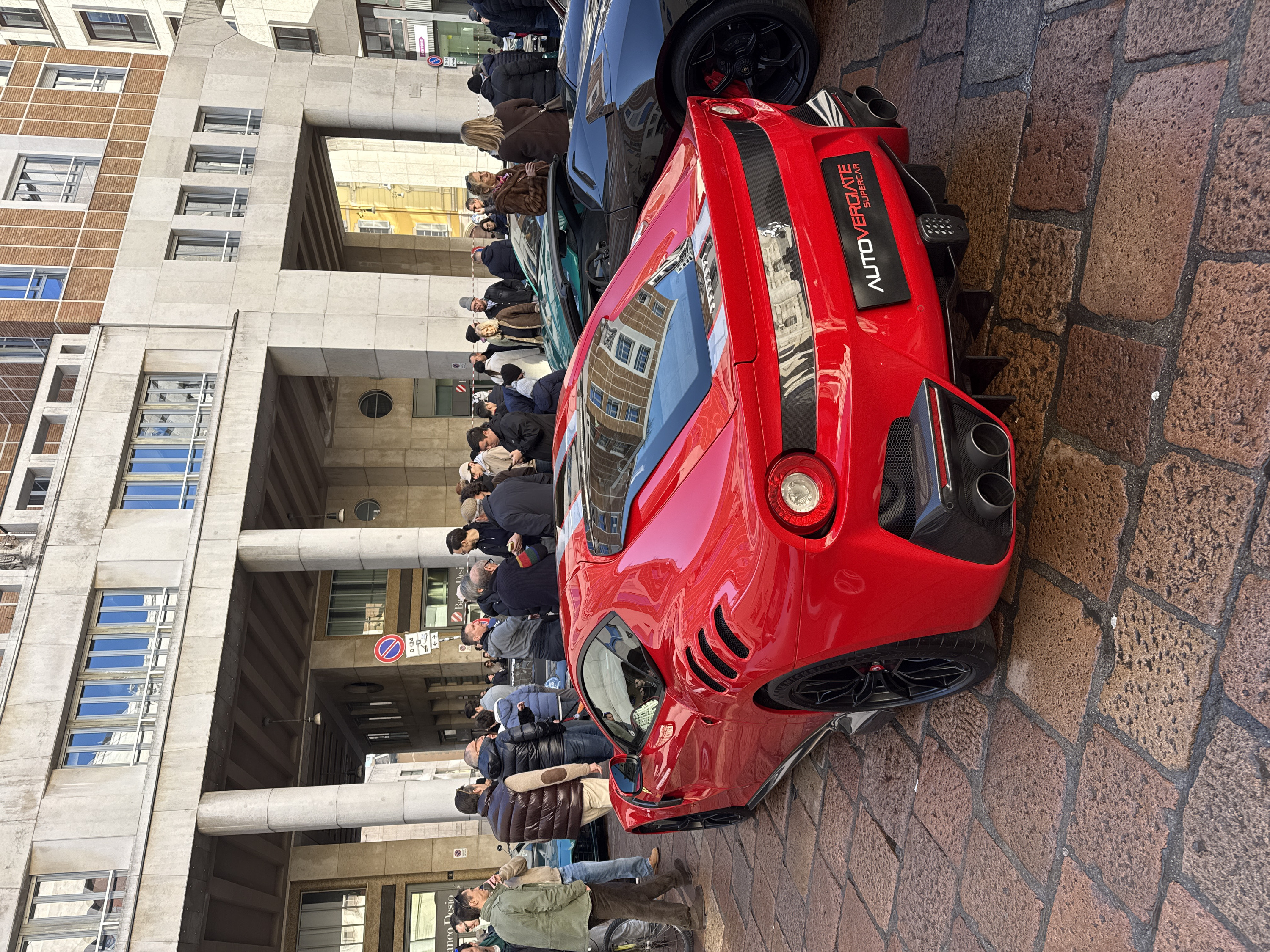 A color photograph captures an outdoor scene on a cobblestone street in an urban environment in Milan, Italy, likely in a central area characterized by both historical and rationalist architecture. The foreground is dominated by the rear three-quarter view of a bright red Ferrari F12tdf supercar, facing away from the viewer. The car features black multi-spoke wheels, a prominent rear diffuser with quad exhaust pipes, and a license plate frame reading "AUTOVERGIATE SUPERCAR."
To the immediate right of the red Ferrari, the front wheel and red brake caliper of a dark grey or black supercar, likely a Lamborghini, are visible. Further behind these vehicles and to their left, a dark green convertible supercar and a partial view of a blue/teal vehicle are present.
Approximately 20-30 individuals, predominantly adults of varying ages, are gathered in groups around the parked supercars. Most are dressed in casual winter attire, including jackets, coats, and puffer vests. Several people are observing the cars, conversing, and some appear to be looking at mobile phones.
The street surface consists of large, irregularly shaped reddish-brown cobblestones. In the background, a multi-story building features a ground-level colonnade with large, smooth concrete pillars supporting upper floors. These upper floors exhibit a mix of light-colored stone or concrete facades with rectangular windows, some of which reflect the sky and surrounding buildings, and sections with a patterned brick texture. Signs for "Banco Desio" and "DENTONs" are visible on buildings, along with a "No Entry" traffic sign and a "P" (parking) sign indicating 24-hour availability. The scene is brightly illuminated by natural daylight.