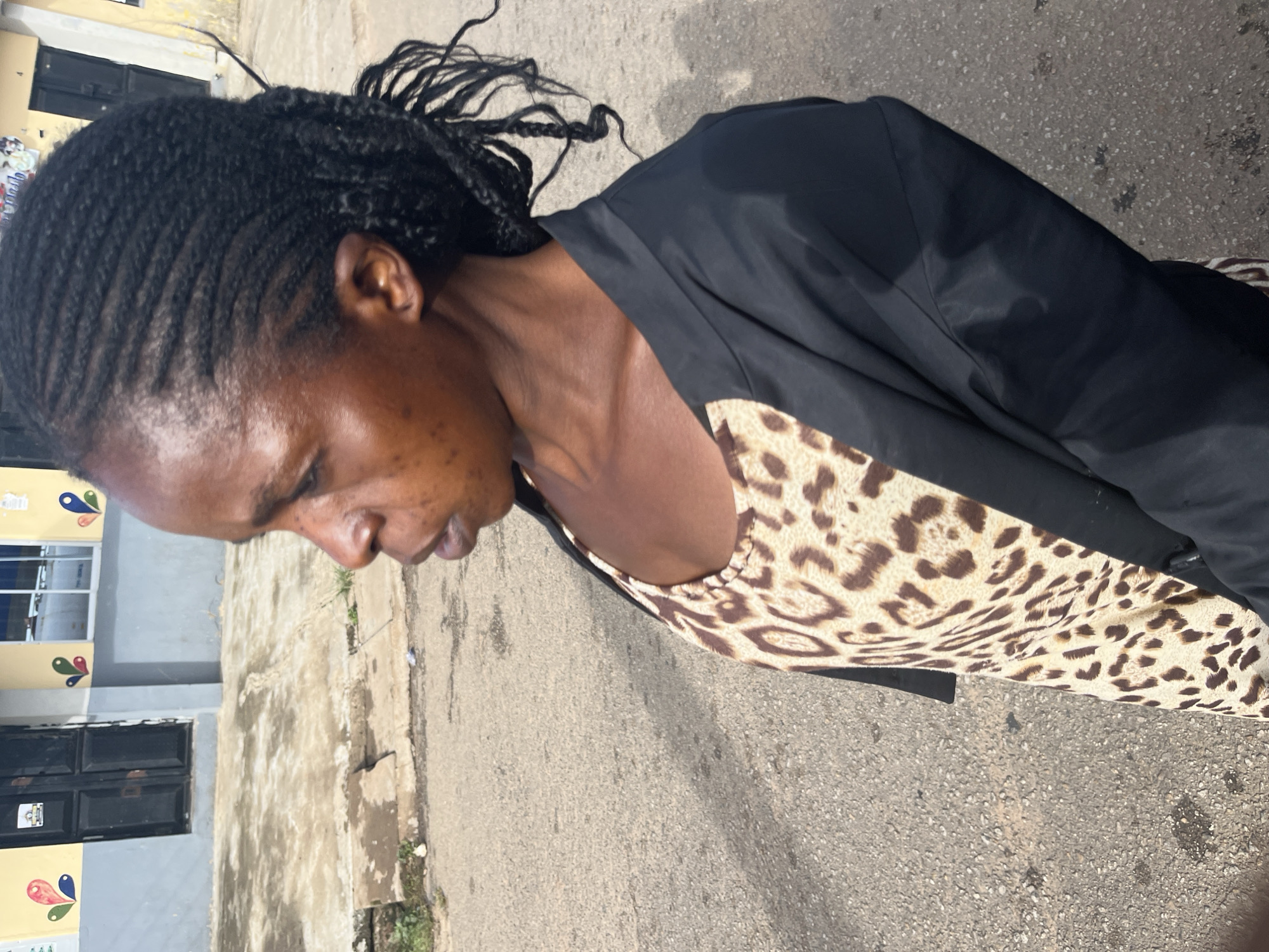 A woman with dark skin and braided hair stands on a paved street in Jos, Nigeria. She is wearing a leopard-print top and a black jacket. The background shows a building with colorful murals on its facade. The street is made of asphalt and shows some signs of wear. The lighting suggests it is daytime.