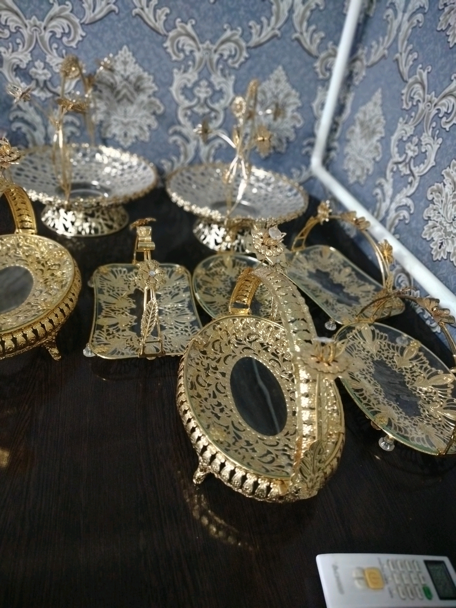 The image displays a collection of ornate, gold-colored serving dishes and trays arranged on a dark, polished surface, likely a table. The items feature intricate filigree designs, floral motifs, and some appear to have glass or mirrored bases. In the background, a textured blue wallpaper with a damask pattern is visible. The scene is brightly lit, creating reflections on the metallic surfaces of the dishes. A white remote control is partially visible in the lower right corner of the image. The context of Көлкент, Kazakhstan is not visually evident in the image itself.