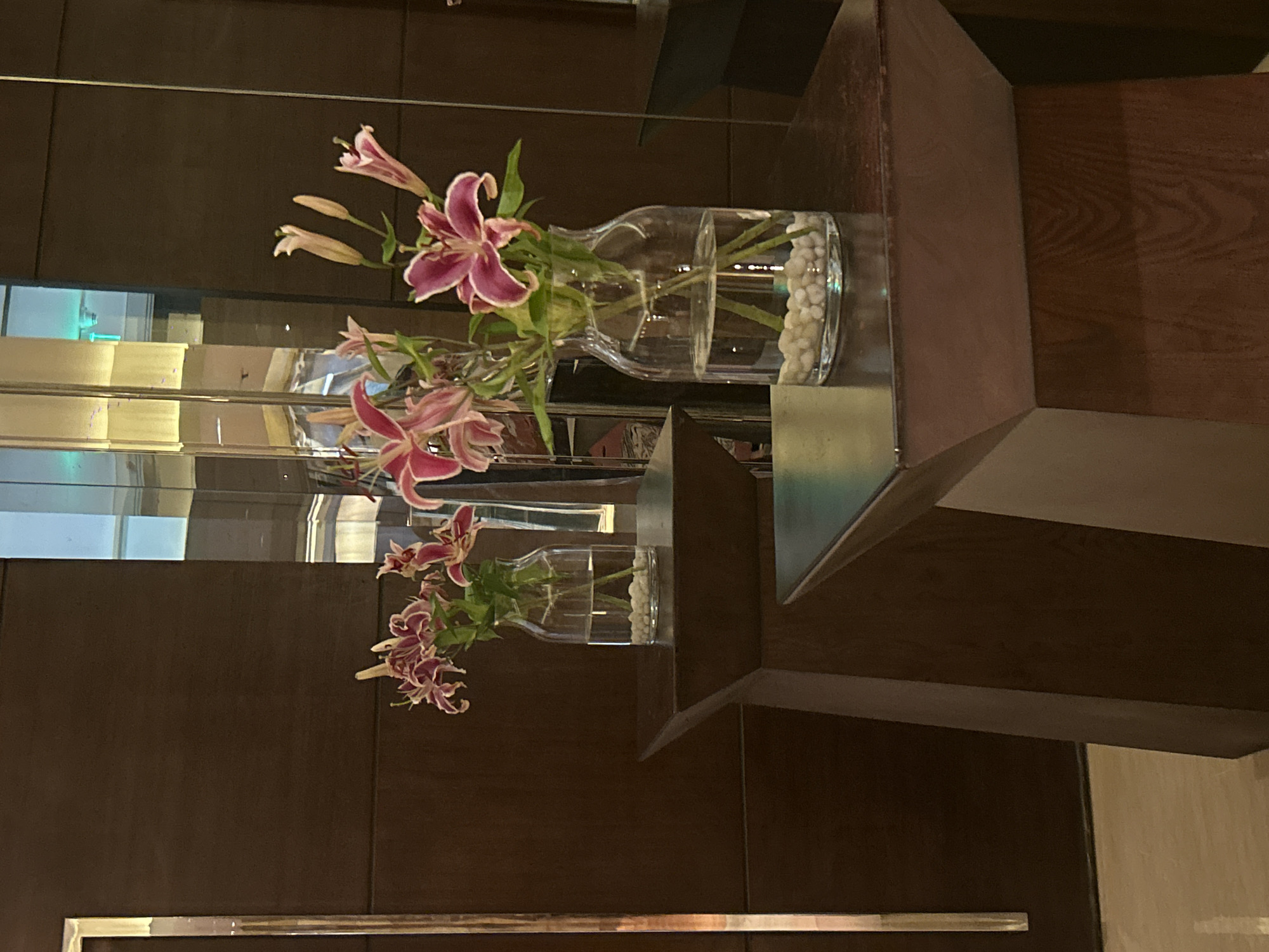 This image depicts two glass vases containing pink lilies and white pebbles, placed on dark wooden pedestals. The pedestals are situated in front of a dark wooden wall with a vertical metallic strip on the left. In the background, a reflective surface, possibly a mirror or glass partition, shows a glimpse of a room with ambient lighting and what appears to be a ceiling feature with green lights. The overall scene suggests an indoor setting, likely a hotel lobby or a well-appointed common area. There are no people visible in the image. The lighting is subdued, with reflections creating highlights and adding depth to the scene. The focus is on the floral arrangements and the architectural elements of the space. The image does not provide any specific information to suggest its location in Pune, India.