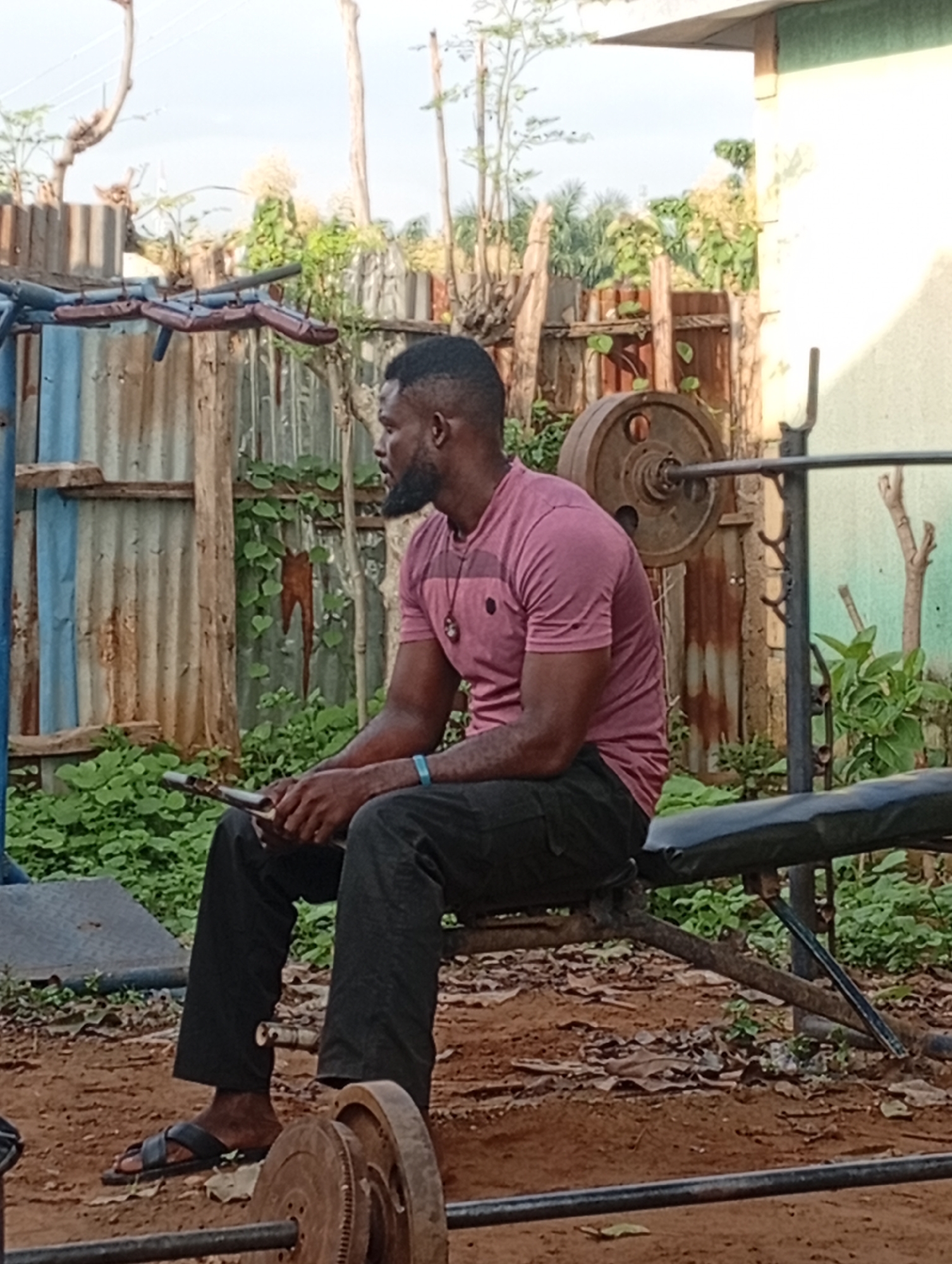 A Black man with a beard sits on a weight bench outdoors, holding a mobile phone in his hands. He is wearing a pink t-shirt and dark pants. To his left, a weightlifting bar with a large weight plate is visible. The background consists of a rustic fence made of corrugated metal and wood, with lush green foliage and trees. A light-colored building is partially visible on the right. The ground is covered with dirt and fallen leaves. The scene appears to be in a residential outdoor area, possibly a makeshift gym, in Akwanga, Nigeria.