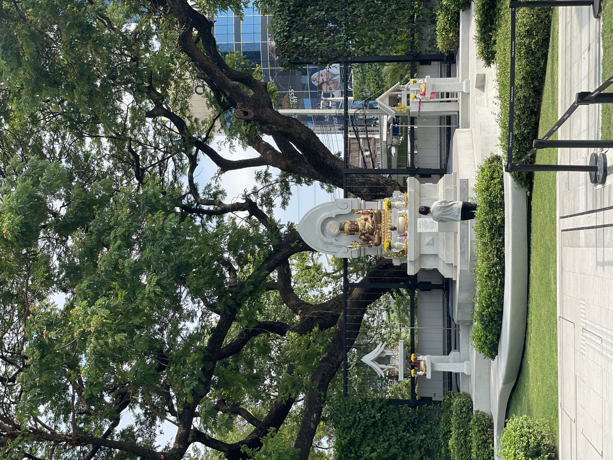 The image captures a Hindu shrine in Bangkok, Thailand. The shrine, made of white concrete, features a golden statue of a deity. A person is praying in front of the shrine. Smaller spirit houses, also white, flank the main structure. Lush greenery, including a large tree with dense foliage, surrounds the shrine. In the background, a modern high-rise building with a large billboard is visible. The setting appears to be a well-maintained public space, possibly a park or a designated religious area.