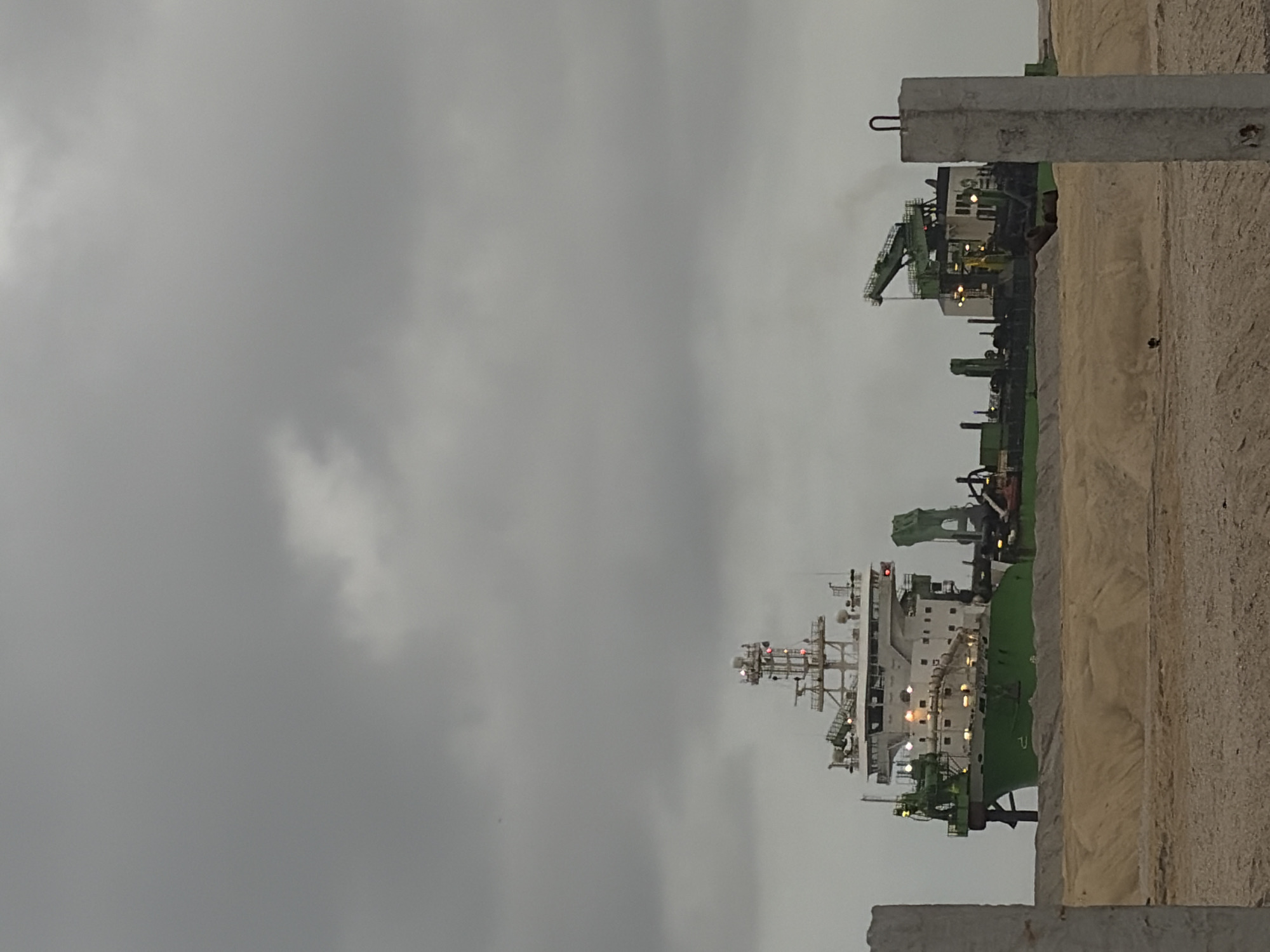 The image shows a large green and white dredging vessel docked in the city of Ilado, Nigeria. The vessel is equipped with cranes and other machinery, suggesting it is actively engaged in dredging operations. The foreground features a sandy embankment, possibly part of the dredging work or a construction site. The sky is overcast, with grey clouds obscuring the sun. Two concrete posts are visible in the foreground, one on the left and one on the right, framing the scene. There are no people clearly visible in the image. The overall atmosphere is one of industrial activity against a somewhat subdued natural environment.