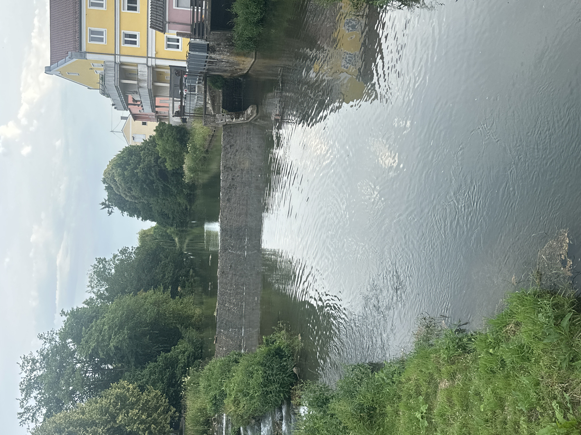The image captures a serene view of the Abens river in Mainburg, Germany. The river flows calmly, with a small weir creating a gentle cascade on the left side, bordered by lush green grass and vegetation. On the right bank, a multi-story building with a distinctive yellow facade stands prominently, featuring balconies and white-framed windows. Adjacent to it, a darker pink building is partially visible. The river's surface reflects the sky, trees, and buildings, creating a tranquil, almost ethereal atmosphere. The sky is overcast, with soft clouds suggesting a mild, perhaps early evening, setting. The scene is devoid of any visible people or animals, emphasizing the natural beauty and peacefulness of the location.