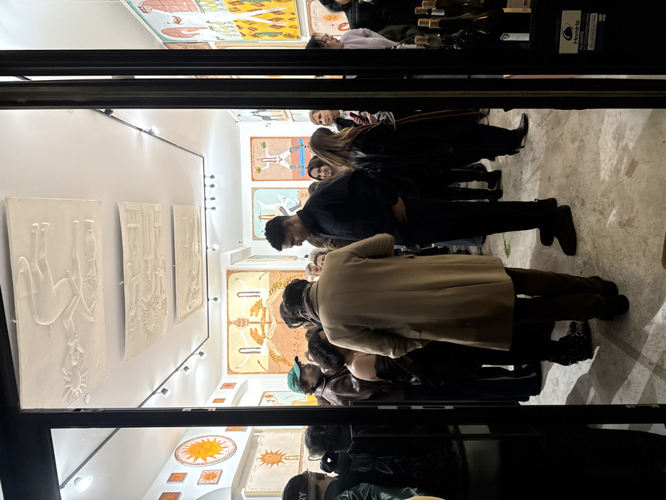 A group of people gathers inside an art gallery. White walls display a mix of framed artwork, including flat paintings with surreal themes and three-dimensional, embossed pieces. The art features depictions of figures, suns, and abstract patterns. The gallery is illuminated by overhead track lighting. The view is partially obstructed by the gallery's entrance.