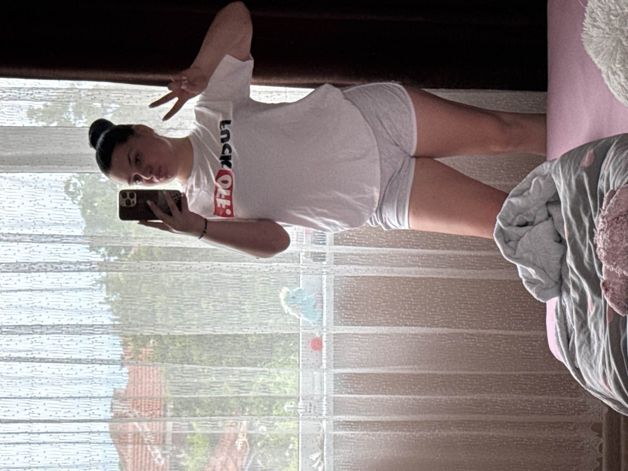 A young woman with dark hair in a bun is standing in front of a window, taking a selfie with her phone. She is wearing a white t-shirt with "FUCK OFF" written on it in red and black lettering, and grey shorts. Her left hand is holding the phone, and her right hand is raised in a peace sign. The background shows a slightly blurred view of trees and a building with a red roof, suggesting an outdoor setting. Inside the room, a bed with pink and grey bedding is visible in the foreground, and a light-colored curtain hangs by the window. A small blue stuffed animal and a red ball are on the windowsill. The scene appears to be an indoor residential space in Rogosh, Bulgaria.