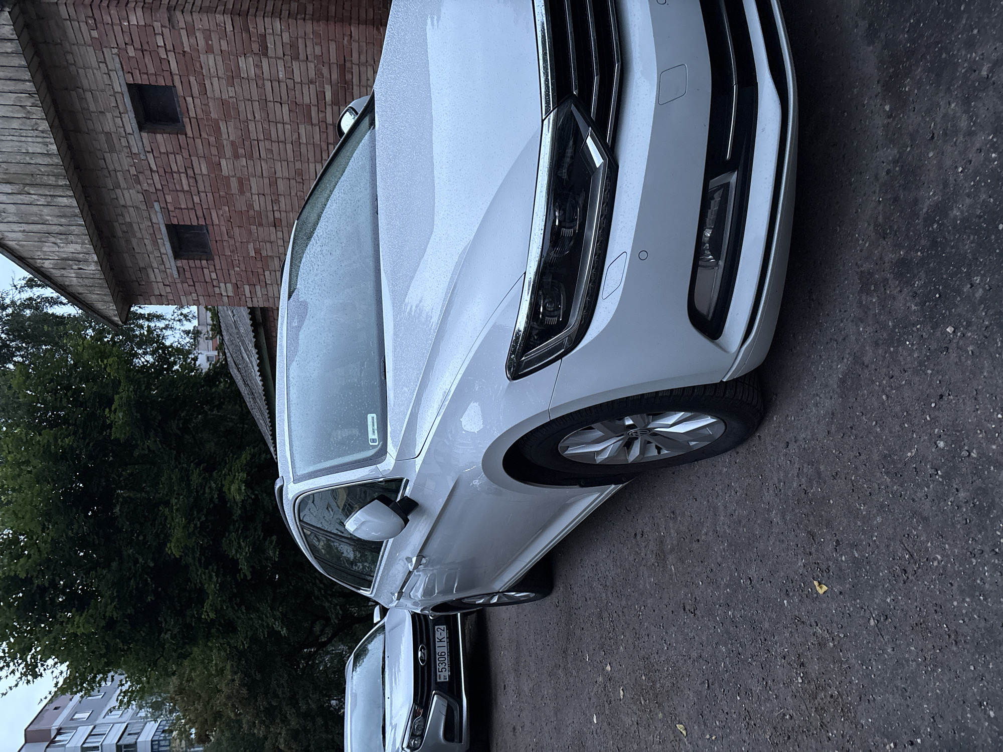 The image shows the front quarter panel, headlight, and wheel of a white Volkswagen Passat parked on an asphalt surface. The car appears wet, suggesting it has recently rained or is being washed. Behind the car, a red brick building with two dark square openings is visible, alongside a wooden overhang. A large tree with dense green foliage occupies the background. In the distance, a multi-story building with numerous windows can be seen. Another car, with a license plate reading "5306 IK-2," is parked in front of the Volkswagen. The lighting suggests it is daytime, possibly overcast due to the wetness of the car and the general subdued light. The scene is likely a street or parking area in Vitsebsk, Belarus. There are no people visible in the image.