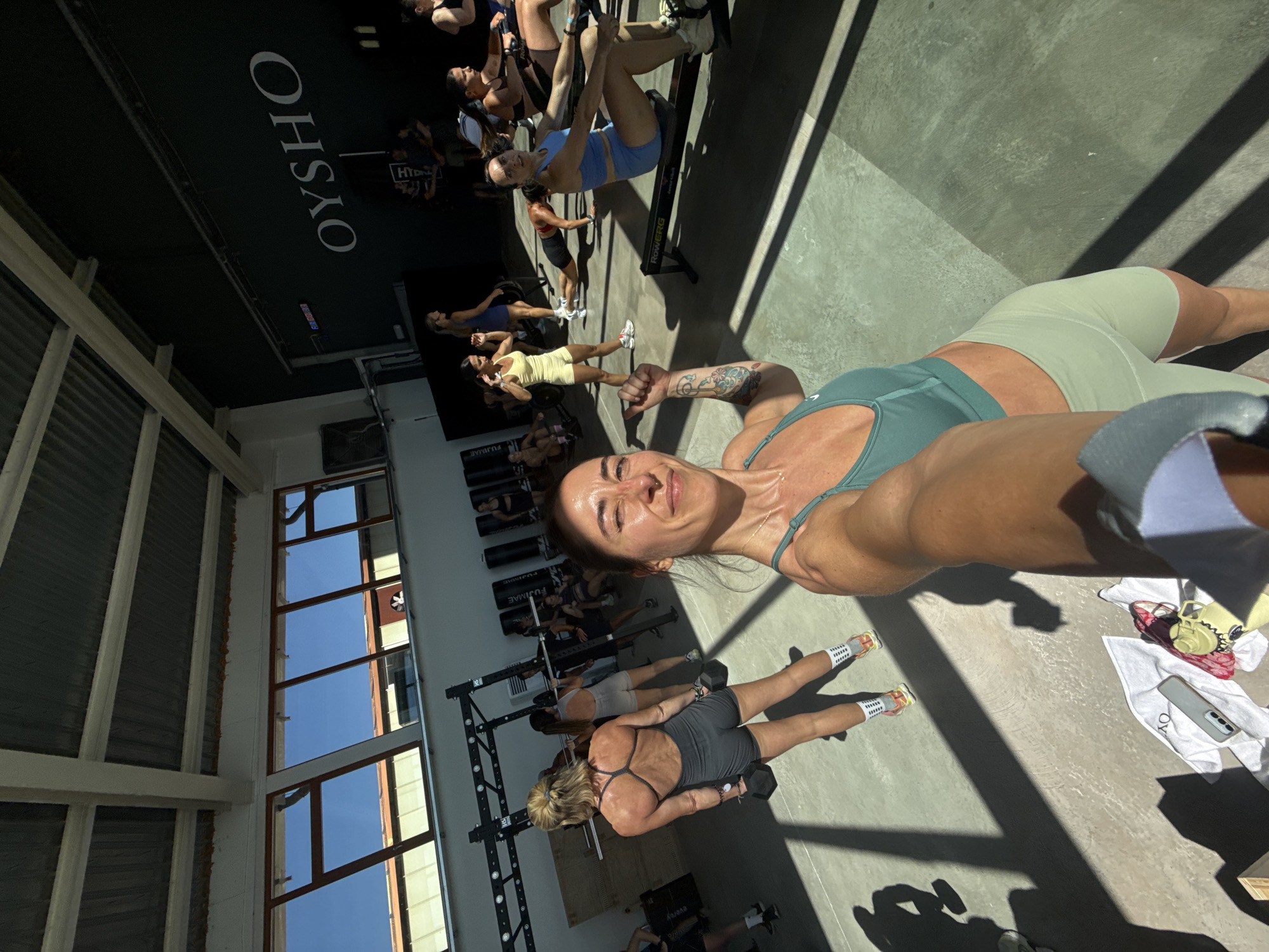 The image depicts an indoor gym environment, likely in Barcelona, Spain, as indicated by the "OYSHO" branding visible on the wall, a Spanish retailer. The gym features high ceilings with exposed beams and large windows that allow natural sunlight to stream in, casting distinct shadows on the concrete floor. Several women are engaged in various fitness activities. In the foreground, a woman takes a selfie, smiling at the camera, wearing a teal sports bra and light green biker shorts. Behind her, other women are seen lifting weights, using rowing machines, and performing exercises. Equipment such as weight racks, punching bags, and rowing machines are present throughout the gym. The overall atmosphere suggests a dynamic and active fitness class or training session.