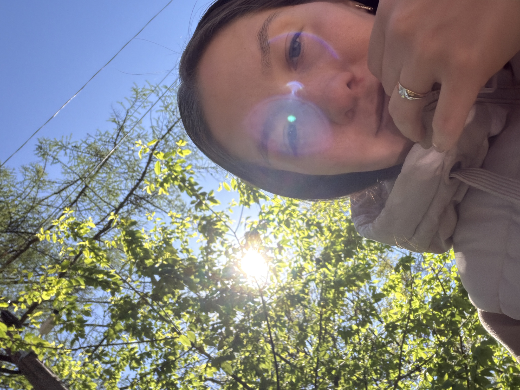 A low-angle selfie photograph shows a person's face, hand, and the upper part of a light-colored, possibly beige or gray jacket. The person's face is centered in the upper portion of the frame, with one hand partially covering the lower part of the face. The hand has a ring with a stone. A bright lens flare obscures part of the face. The background consists of a clear, blue sky visible in the upper-left corner and green foliage, presumably trees, with the sun shining through the leaves. The location is outdoors, likely in a park or wooded area, and not explicitly located in Staraya, Russia.