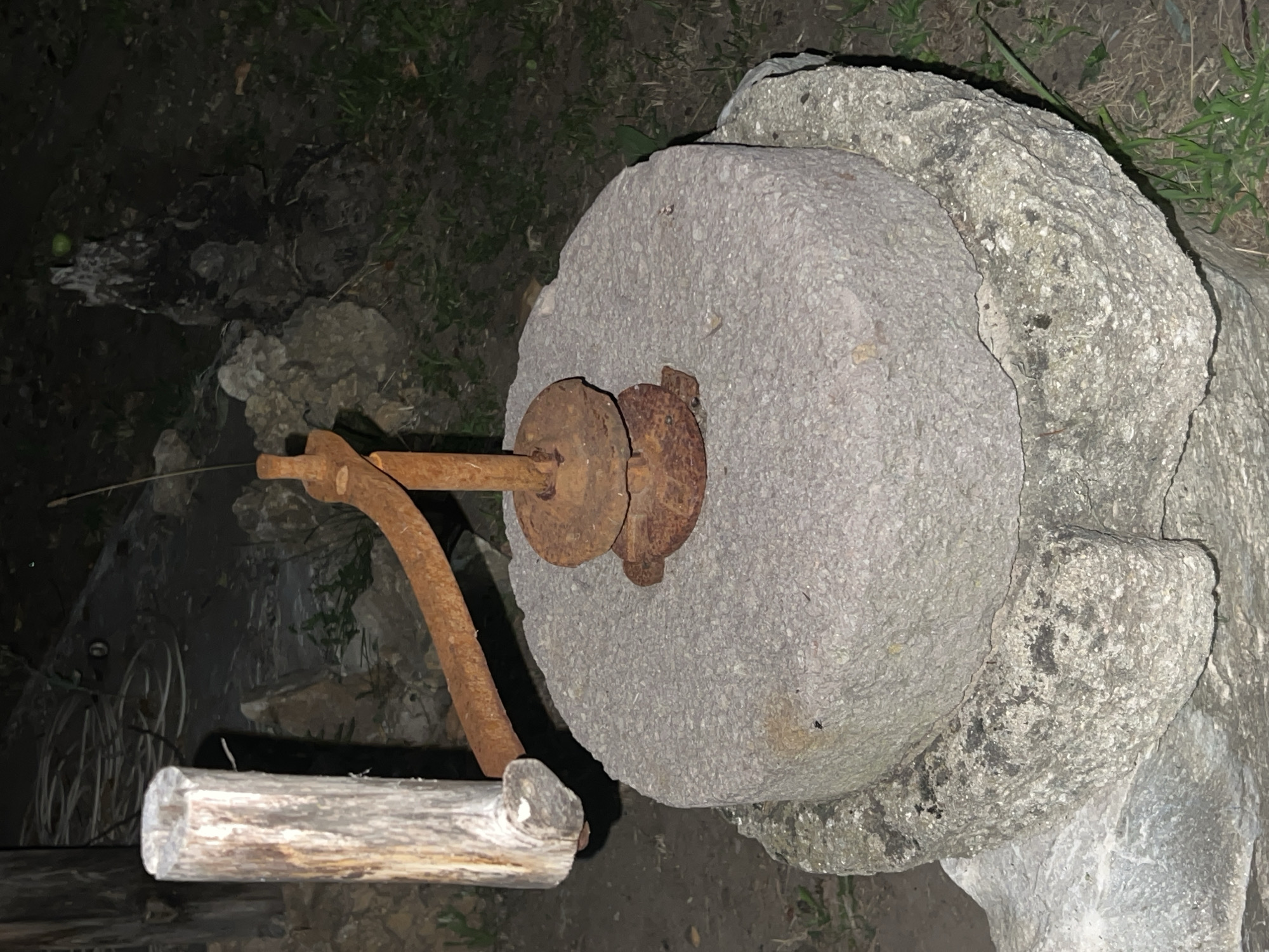 The image shows a close-up of an old, weathered stone mill, likely used for grinding grain. The millstone, made of rough, grey stone, rests on a larger, chipped stone base. A rusty metal mechanism is attached to the top of the millstone, with a wooden handle extending from the left side, suggesting it's a manual grinding tool. The surrounding environment is dimly lit, with visible grass and some dirt or soil. The overall impression is one of age and disuse, hinting at a historical or rustic setting. The location is identified as Akandzhievo, Bulgaria.