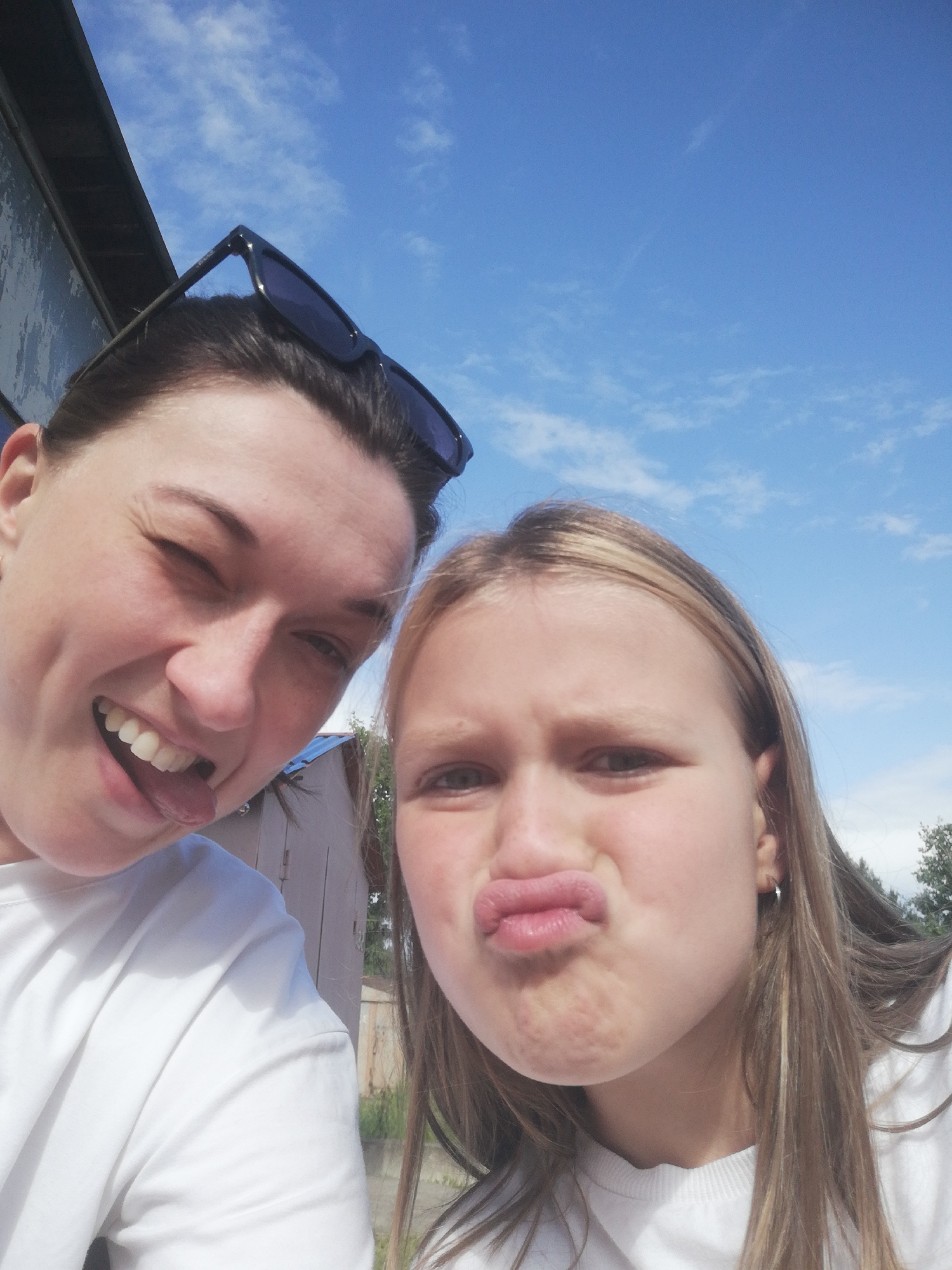 This is a selfie taken outdoors on a sunny day with a blue sky and scattered clouds. Two individuals are visible. The person on the left, closer to the camera, is a woman with dark hair, wearing sunglasses perched on her head. She is smiling broadly with her mouth open and tongue sticking out. The person on the right is a younger individual, likely a girl, with blonde hair. She is puckering her lips for the camera. Both are wearing white shirts. In the background, a light-colored building, possibly a shed or small structure, is visible, along with some greenery and trees. The overall setting suggests a casual, outdoor activity. The image does not contain any specific landmarks or elements that would place it in the city of Tosno, Russia.