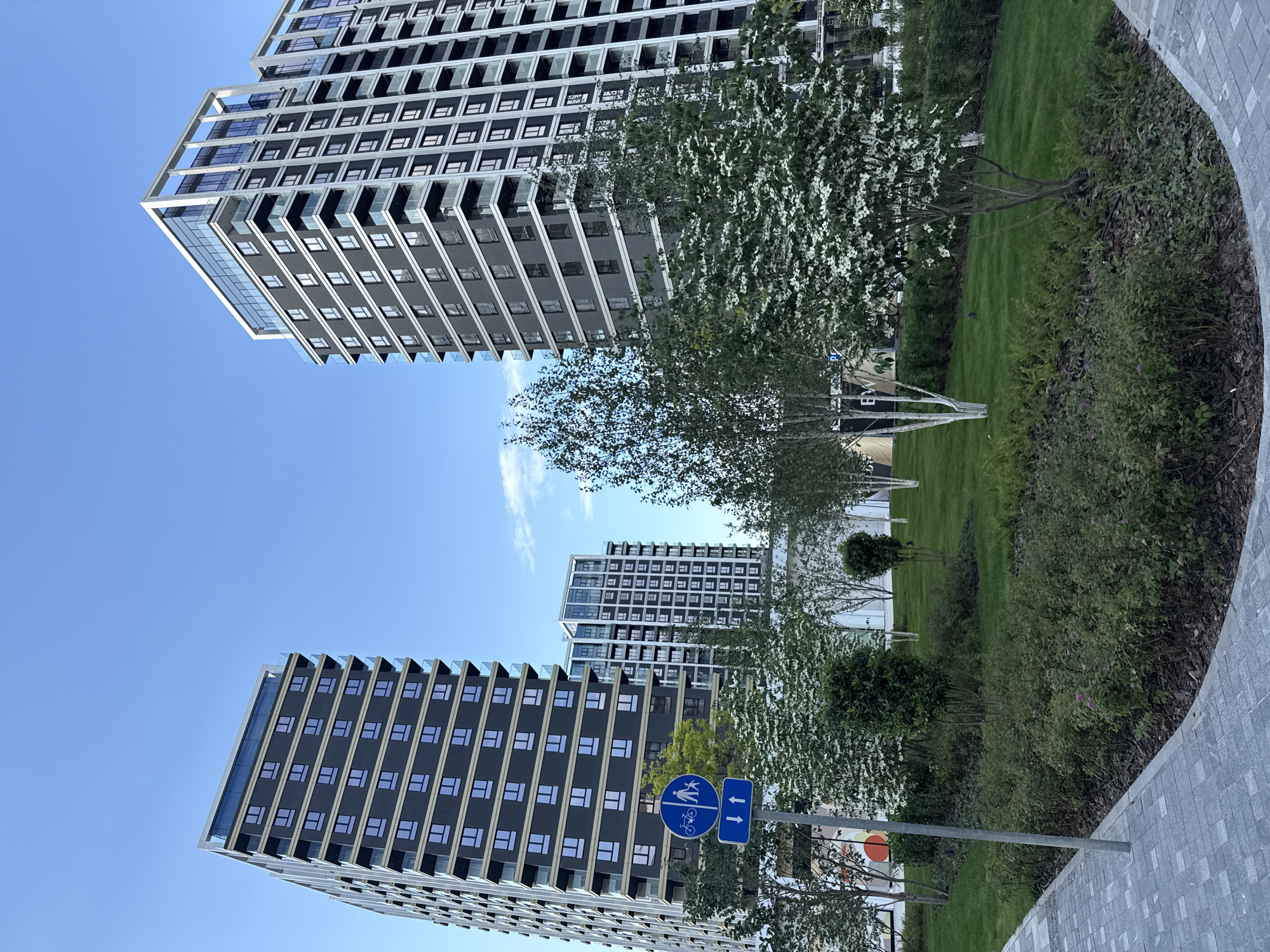 The image shows three modern buildings in Belgrade, Serbia, viewed from a low angle. The buildings are tall and have a repetitive window pattern with prominent balconies. Between the buildings are landscaped areas featuring green grass, shrubs, and trees, some with white blossoms. In the foreground is a pathway made of grey pavers. A sign with a blue background indicates a shared path for pedestrians and cyclists. The sky is blue with a few wispy clouds.