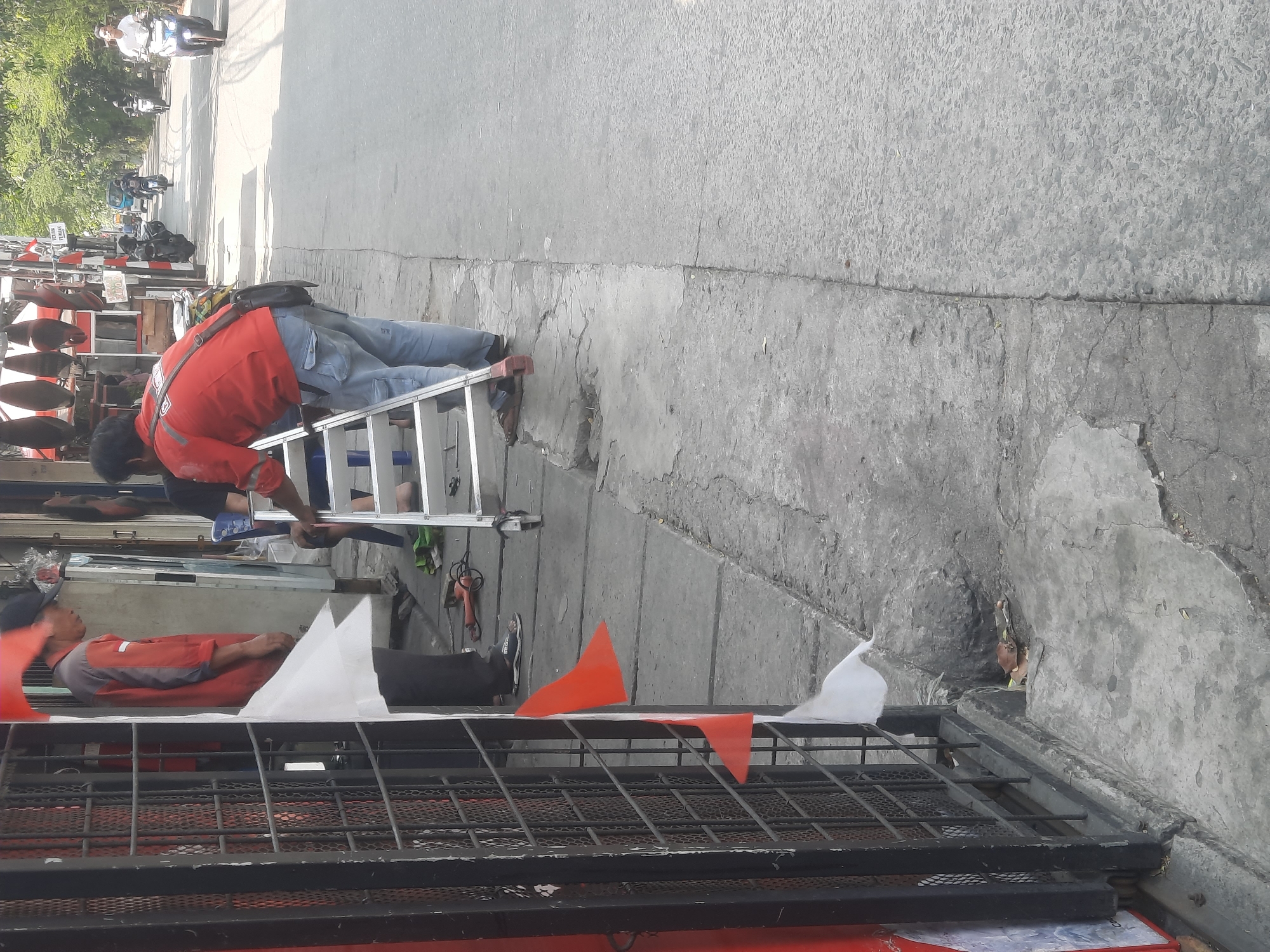 In North Jakarta, Indonesia, a man in a red shirt and blue jeans is standing on a sidewalk next to a street, using a portable aluminum ladder. He appears to be working on something at an elevated position, as his hands are reaching upwards. The ladder is set up perpendicular to the street, with its base on the cracked concrete sidewalk. Behind him, on the same sidewalk, another person in an orange shirt and black cap is standing. Several motorcycles are visible on the street, some parked and others in motion, indicating traffic. The scene is characterized by a commercial area, with shops lining the side of the street and various items displayed outside. Red and white flags are attached to a metal gate on the left side of the image, suggesting a festive or patriotic occasion. The ground is a mix of concrete and asphalt, with some debris and a drain visible on the sidewalk. The overall lighting suggests it is daytime, with shadows cast by the structures and people.