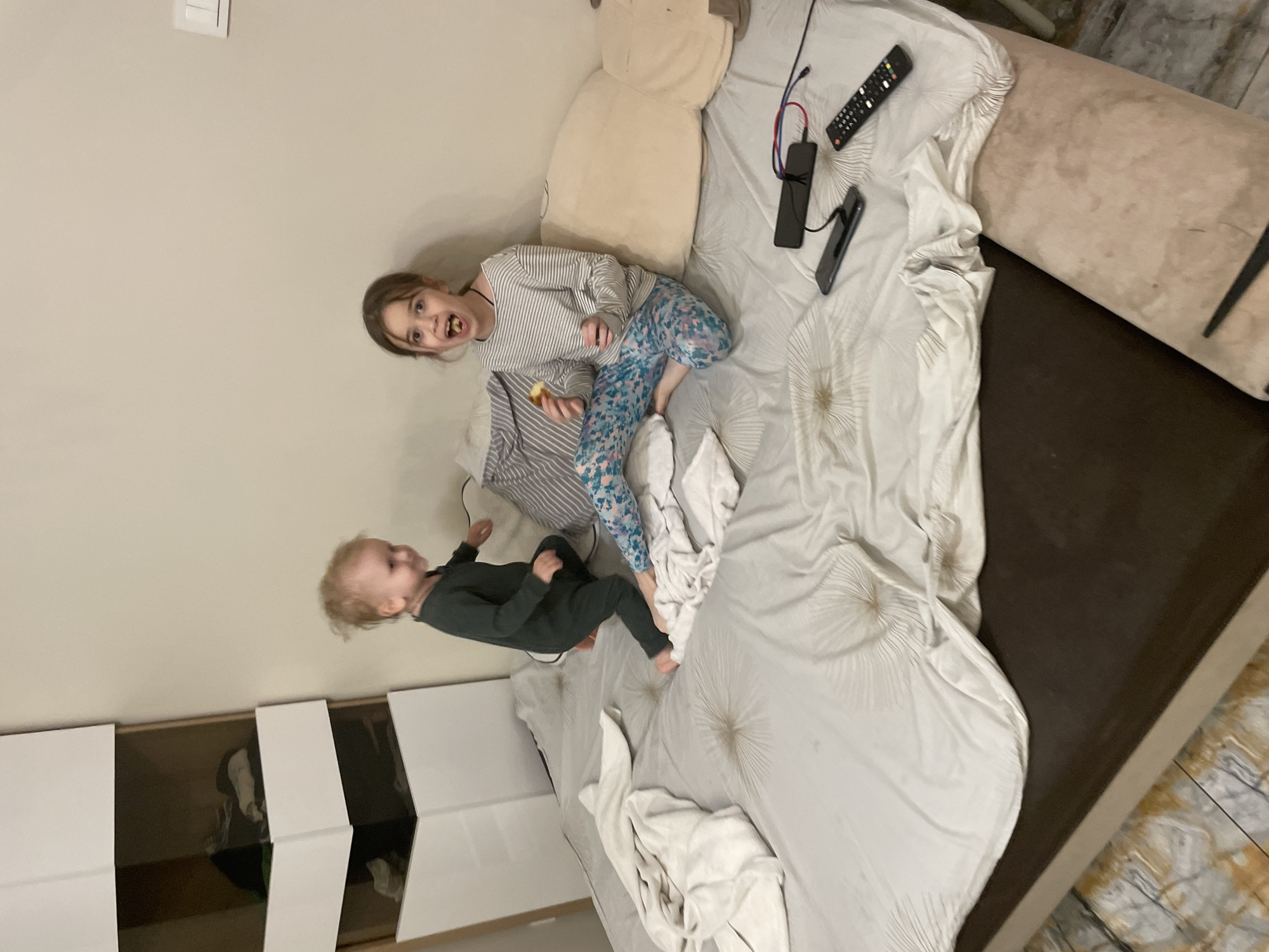 The image shows a room with a light-colored wall on the left. A white dresser with open drawers is visible against the wall. In the center, there is a bed with a brown base and a white comforter. On the bed, there is a girl with brown hair, wearing a striped shirt and patterned leggings. She is holding a piece of food in her hand and has some in her mouth. Next to her, there is a younger child with blonde hair, wearing a dark green outfit. The child is standing on the bed. There are also several objects on the bed including a remote control, a black power bank, and a phone. The location in the city is unknown.