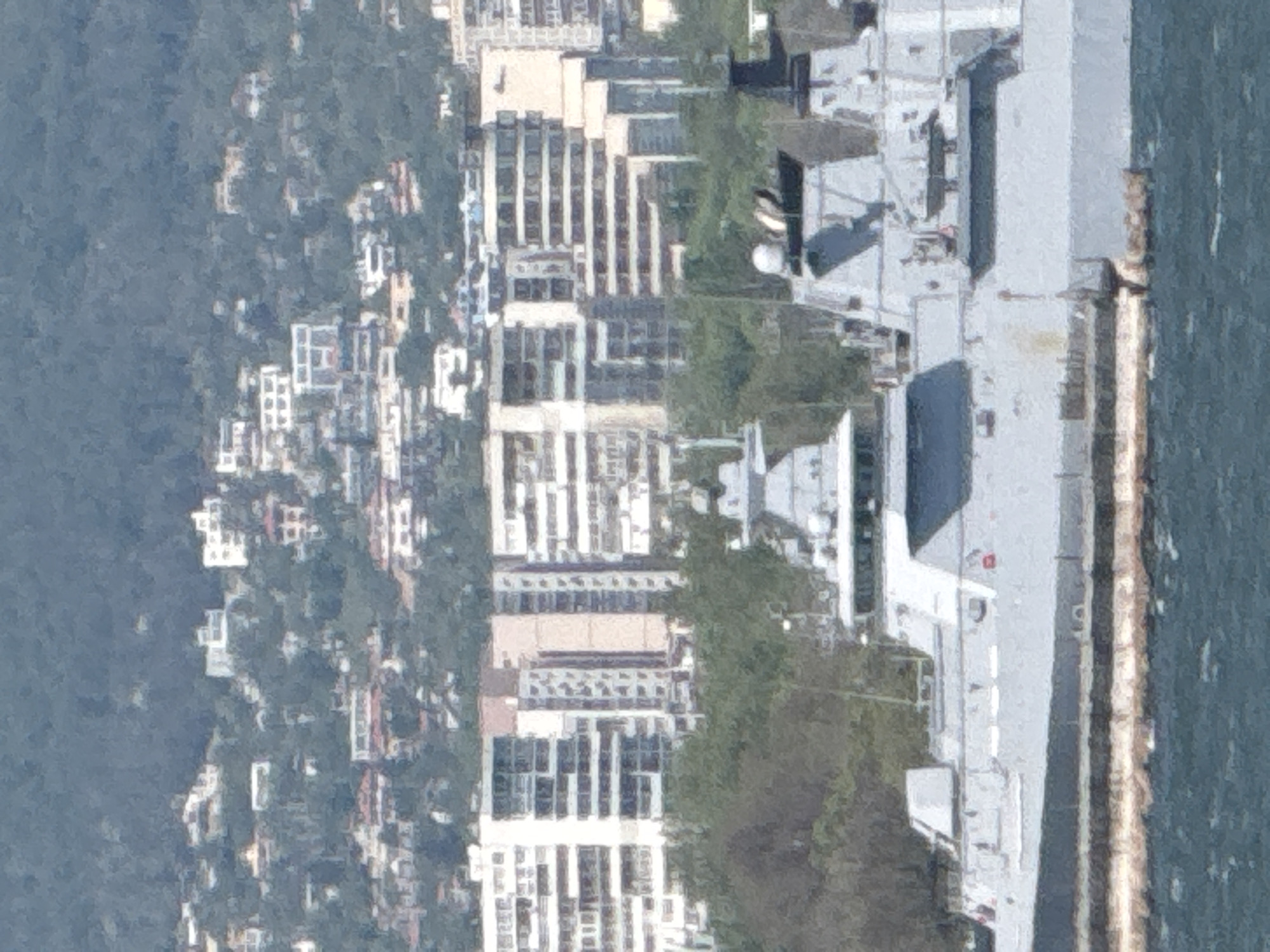 The image shows an aerial view of a cityscape adjacent to a body of water. The buildings appear to be residential or commercial structures, with predominantly white facades and multiple stories. Lush vegetation is interspersed among the buildings. A large, grey ship is docked along the water's edge. The environment is characterized by a blend of urban development and natural elements, with a possible coastline visible in the background. Based on the presence of specific buildings and the layout of the urban area alongside the water, it is very likely that this image shows the city of Varna, Bulgaria.