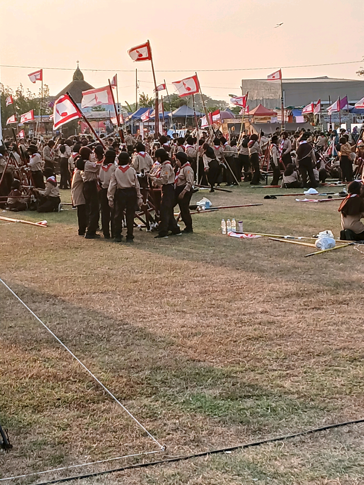 A wide shot captures a large gathering of scouts, possibly during an event or ceremony, on a grassy field under a pale sky. The scouts are dressed in uniforms, many wearing khaki shirts and dark pants, with some also wearing neckerchiefs. Many are holding flags, the majority of which are red and white with a white emblem on a red field. In the background, tents and buildings are visible, including a building with a domed roof, possibly a religious structure. The ground is covered in dry grass, and some areas have visible white ropes or guidelines. There are also several water bottles and other items scattered on the grass. The overall impression is one of an outdoor activity or festival. The image appears to be taken at ground level, looking towards the gathering.