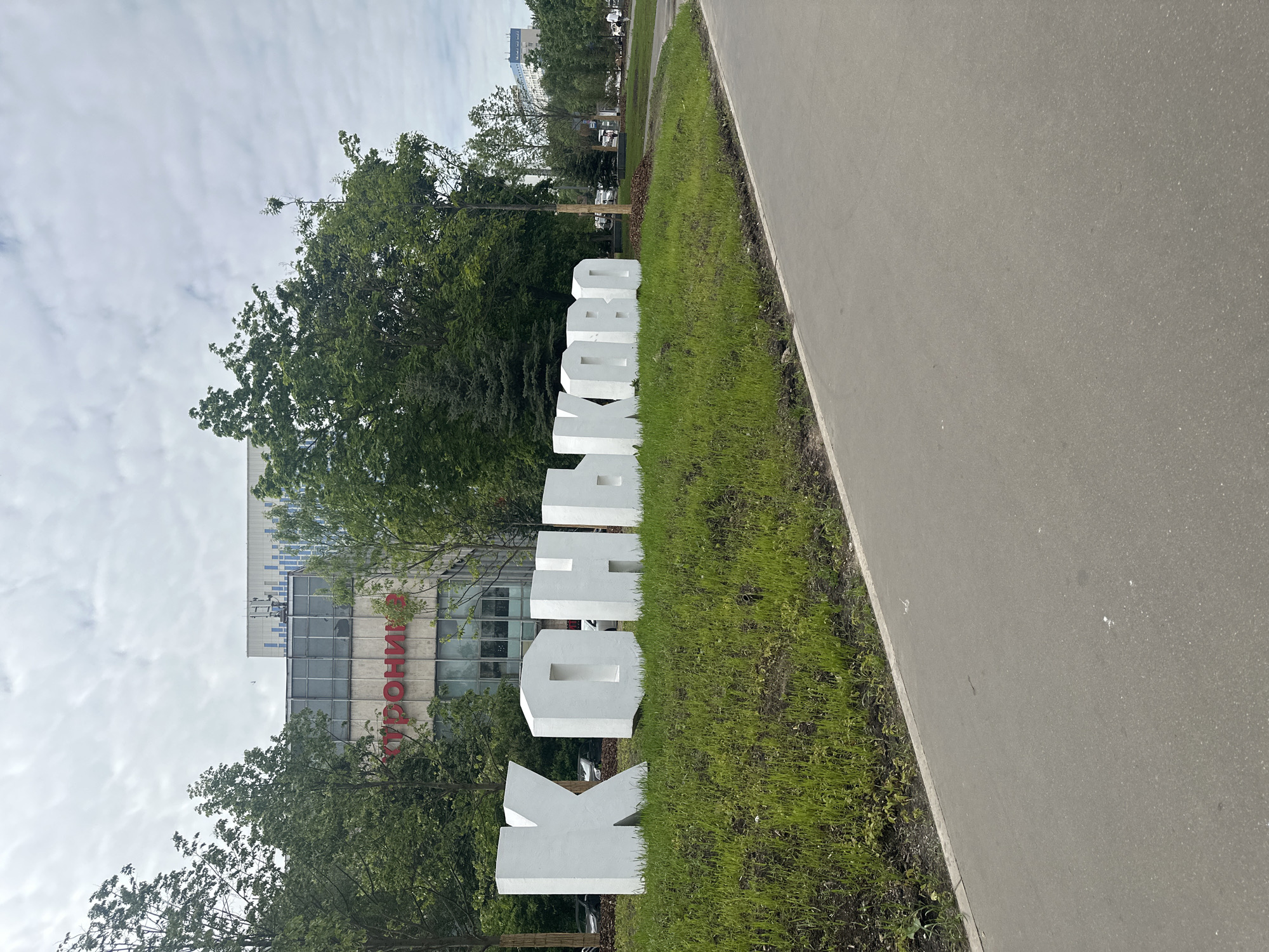 The image, rotated 90 degrees counter-clockwise, displays a large outdoor installation of white, three-dimensional Cyrillic capital letters spelling "КОСМОС" (KOSMOS). These letters are positioned on a strip of green grass. Adjacent to the grassy area, on the right side of the frame, is a paved asphalt path or road.
Behind the letter installation and grass are numerous green trees, their dense foliage indicating a lush environment. In the background, partially obscured by the trees, stands a modern building with a glass facade. Red Cyrillic letters are visible on this building, clearly showing "РОНИЯ" which appears to be part of a larger word such as "УРОНИЯ".
The sky above is overcast with light, wispy clouds, suggesting a daytime setting. There are no people visible, and no specific activities are taking place, presenting a tranquil scene of a public outdoor space, likely in Moscow, Russia.