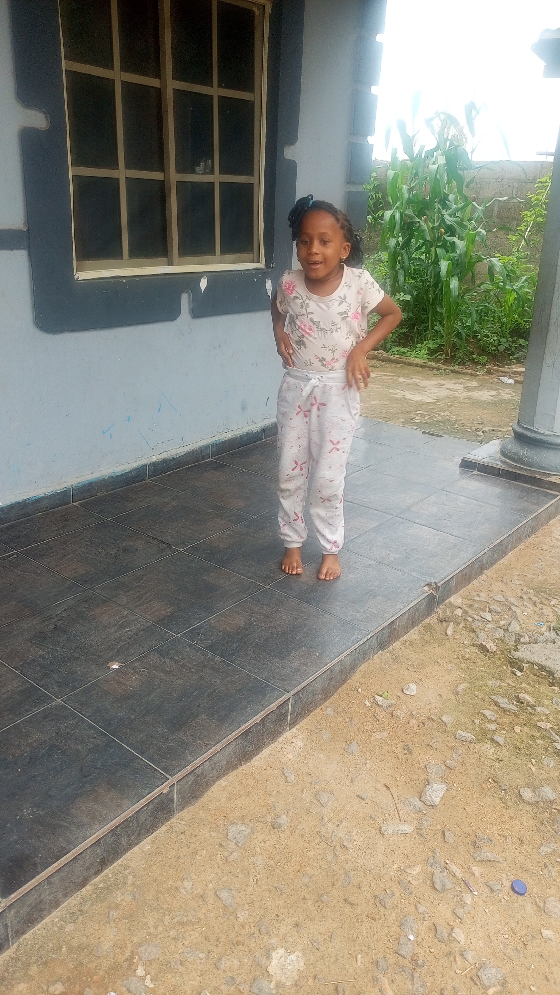 A young Black girl with a black hair ribbon stands on a tiled porch outside a light blue building with a dark gray window frame. She is wearing a white t-shirt with a floral pattern and white pants with a similar floral pattern. Her hands are on her hips, and she is looking at the camera. Behind her, to the right, is a patch of tall green plants, possibly corn. The ground in front of the porch is covered in sand and small rocks. The overall scene appears to be in a residential outdoor setting, possibly in a rural or suburban area.