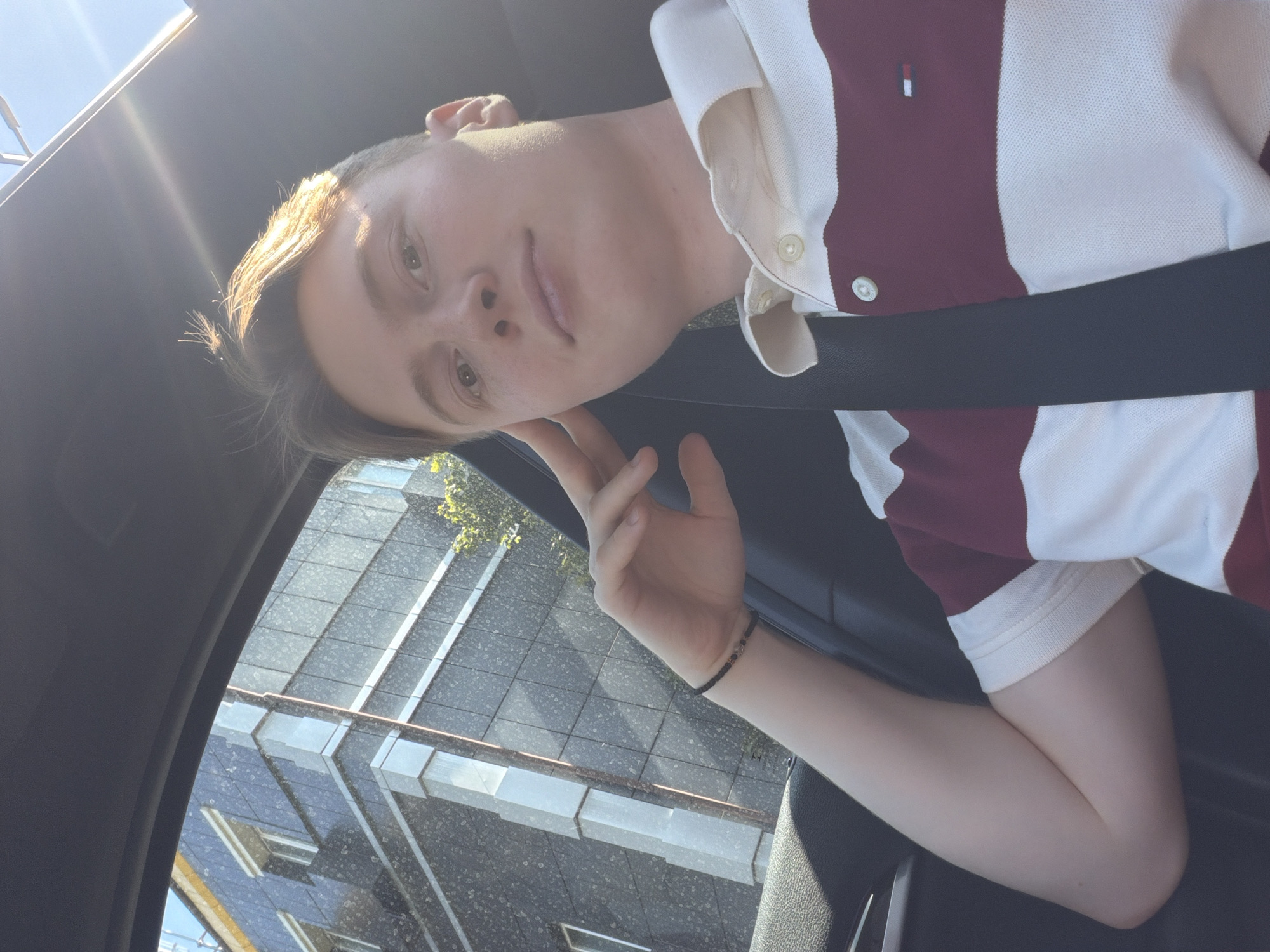The image is a selfie taken from inside a car, looking out towards a modern building in Kyiv, Ukraine. The building has a dark facade with horizontal white accents and square windows. Sunlight illuminates the scene, creating lens flare and highlighting the subject's hair. The subject, a young person with fair skin and light brown hair, is wearing a collared shirt with horizontal burgundy and white stripes. They are resting their head on their hand, with their arm bent and resting on the car's interior. A black seatbelt is visible across their chest. A beaded bracelet is worn on their left wrist. The overall impression is a casual moment captured during the day.