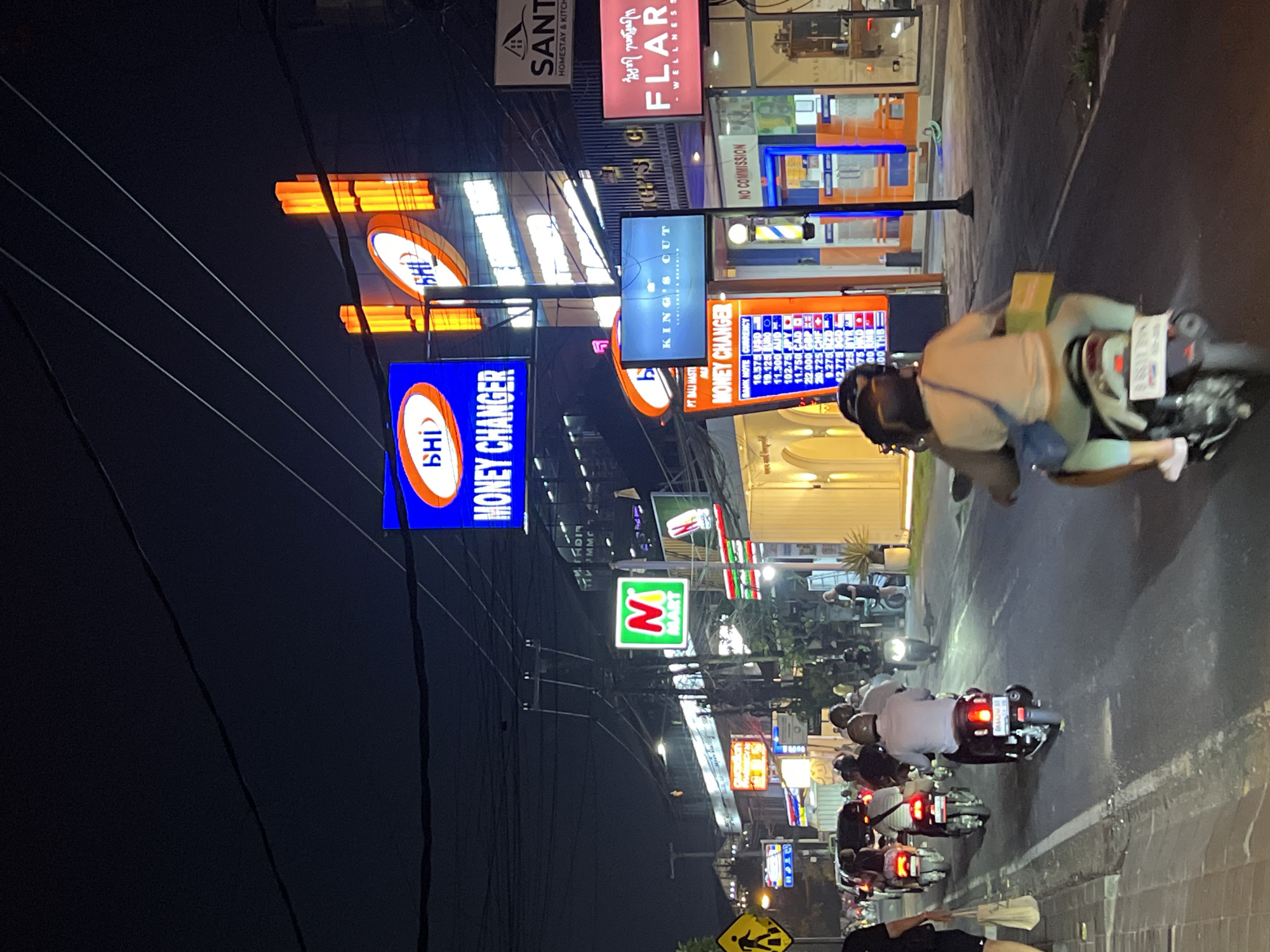 The image depicts a night scene on a street in Canggu, Indonesia, with numerous scooters driving on the right side of the road. The street is lined with businesses, many of which are identified by illuminated signs. Signs include "BHI Money Changer," "King's Cut," "N Mart," "Santi Homestay & Kitchen," and "Flare Wellness." The image shows a money exchange rate board as well. People are seen riding the scooters, and there are electrical wires crisscrossing above the street. The overall environment is urban and bustling, likely a commercial area.