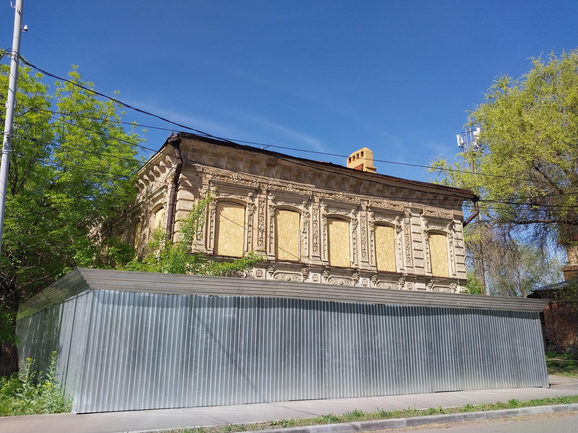 The image depicts a two-story, possibly historical building located in Orenburg, Russia. The windows of the building are boarded up with light-colored wood panels. Above the windows, the building features decorative architectural details. A corrugated metal fence or enclosure stands in front of the building, obscuring the lower portion of the facade. The sky is clear blue. Green trees surround the building. A utility pole is visible on the left side of the frame, with electrical wires running across the top portion of the image. A chimney is visible on the roof of the building. A brick building is visible on the right side of the image. The sidewalk is gray.