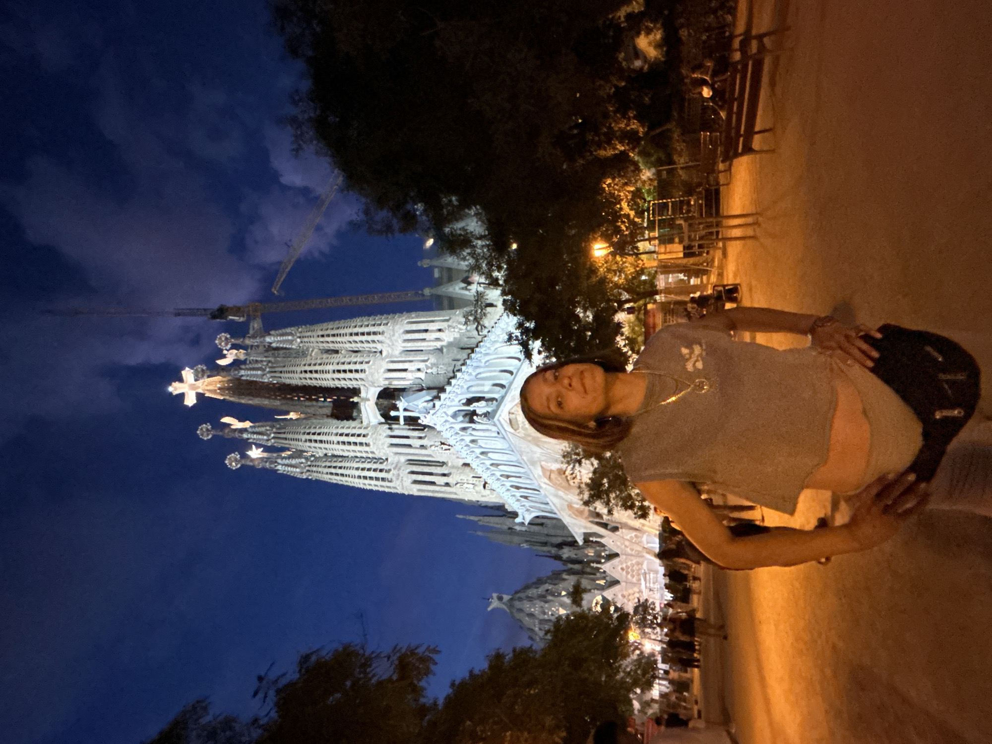The image shows a woman standing in front of the Sagrada Familia in Barcelona, Spain. The Sagrada Familia is a large, unfinished Roman Catholic minor basilica. The woman is wearing a gray crop top, gray shorts, and a fanny pack around her waist. She is standing in a park-like area with trees and benches. In the background, there are construction cranes on the Sagrada Familia. The sky is dark blue, indicating it is nighttime. There are other people in the background, walking around and sitting on benches. The lighting in the park is orange and warm.