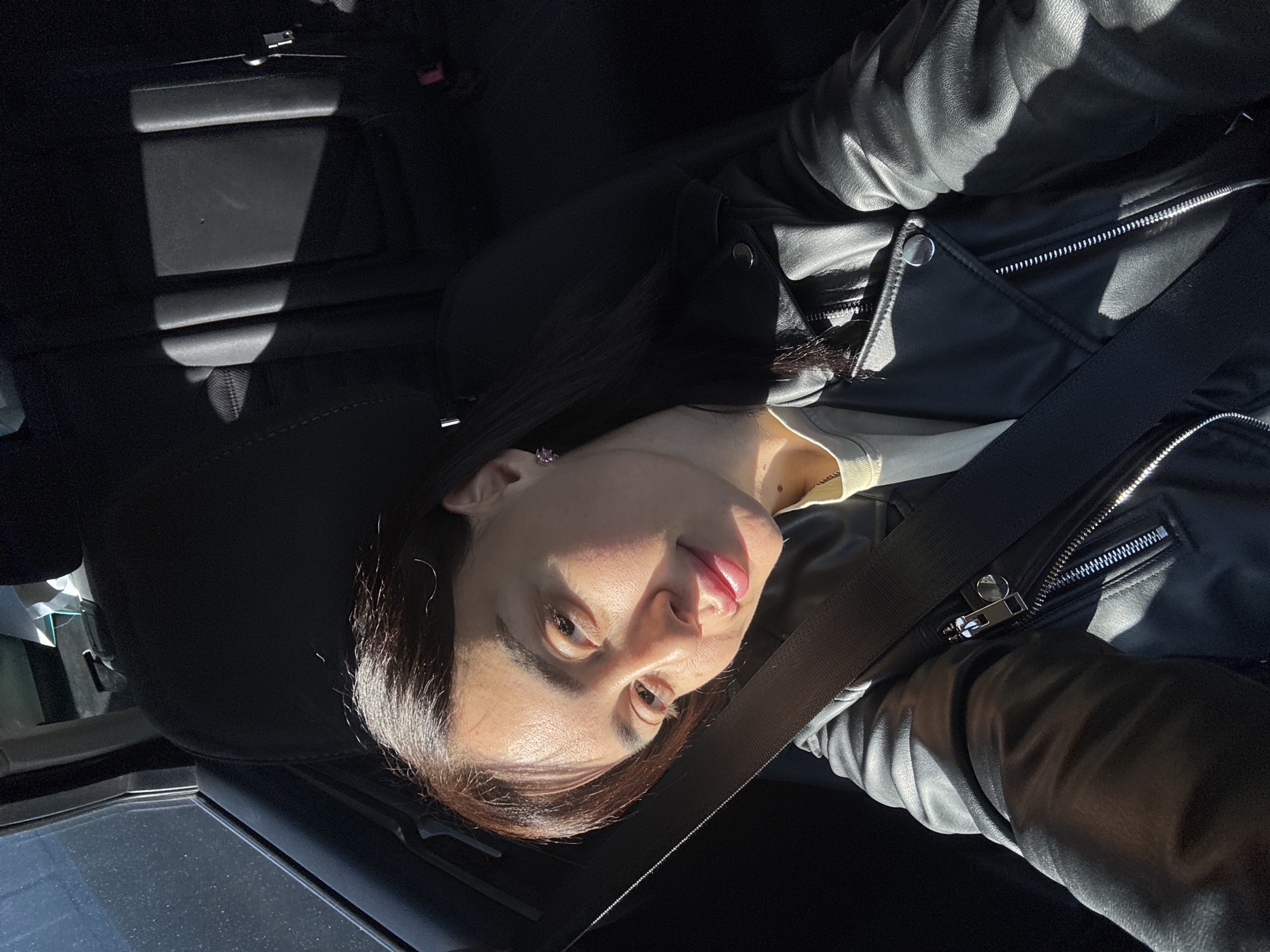 A person is seated in the passenger seat of a car. They are wearing a black leather jacket and a seatbelt. The interior of the car is visible, with black seats and a window. The location is unknown and cannot be determined from the image.
