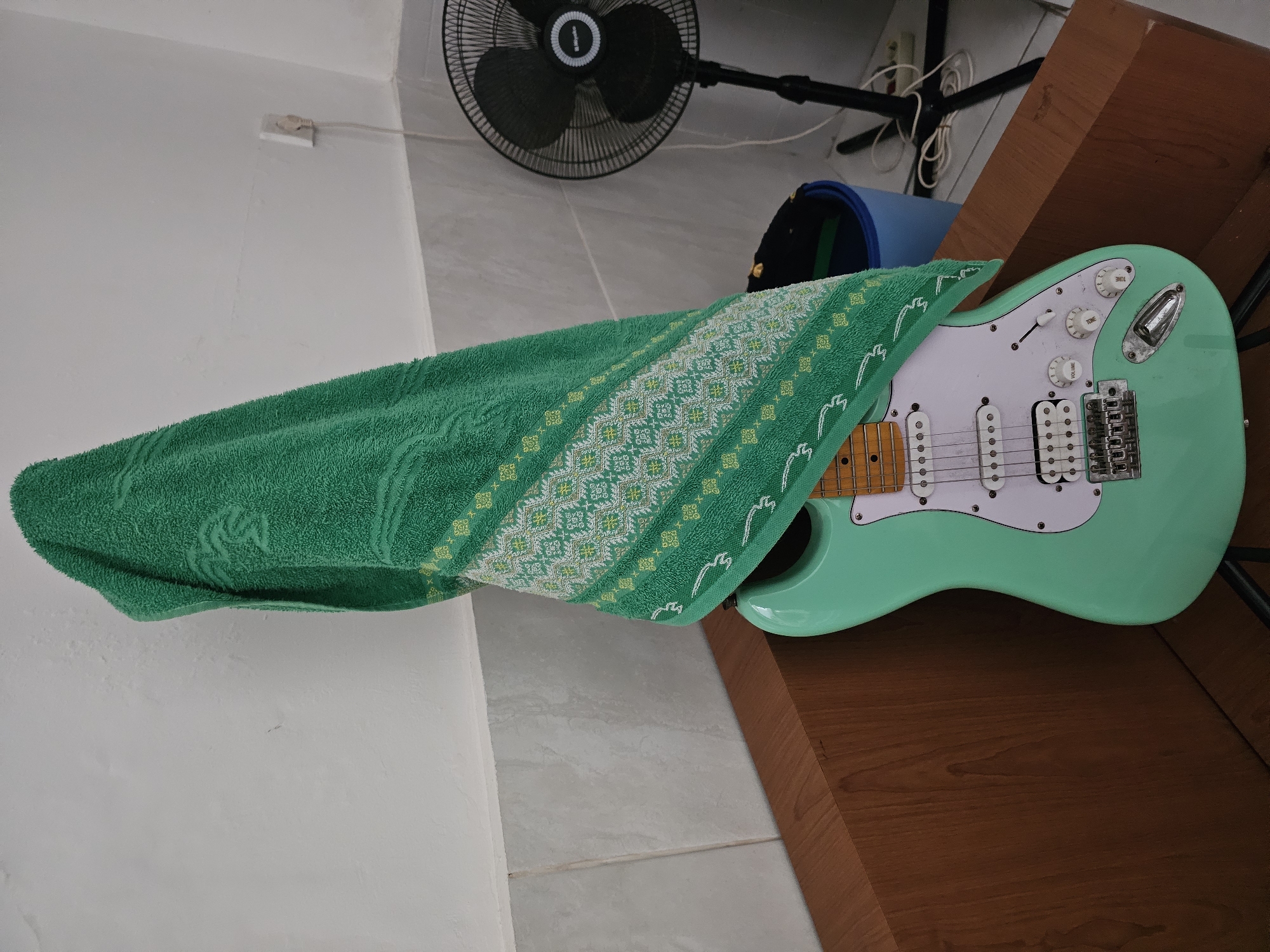 A light green electric guitar with a white pickguard and chrome hardware is covered partially by a green towel with embroidered designs, resting on a brown wooden surface. Behind the guitar, a black stand fan and a blue bag are visible. The background includes a white wall with an electrical outlet. This scene appears to be indoors. I do not have the information required to determine if the scene is within the city of Denpasar, Indonesia.