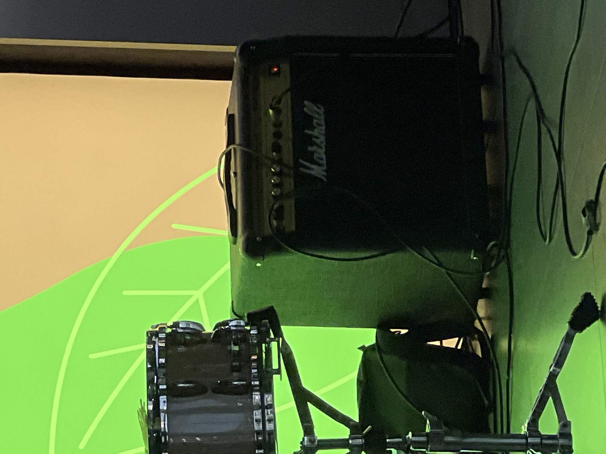 This image shows a close-up of a Marshall amplifier, likely used for musical performances. The amplifier is black with the distinctive white "Marshall" logo visible. There are several knobs and a small red indicator light on the top panel. Cables are connected to the amplifier, suggesting it is in use or set up for a performance.
In the foreground, partially visible, is a drum, possibly a snare drum, mounted on a stand. The drum appears to be made of a dark material with metal accents. Part of a cymbal stand is also visible.
The background features a large screen or wall with a light-colored surface. A large, stylized green leaf design is projected or painted onto this surface. The overall impression is of a stage or performance space, possibly at an event or concert.
The lighting suggests an indoor setting, possibly in the evening or with artificial lighting. The colors are somewhat muted, with a focus on the black of the amplifier and drum, and the green and beige of the background.
No people are visible in the frame. The image is focused on the musical equipment and stage setup. The setting is identified as Ladozhskoe Ozero, Russia.