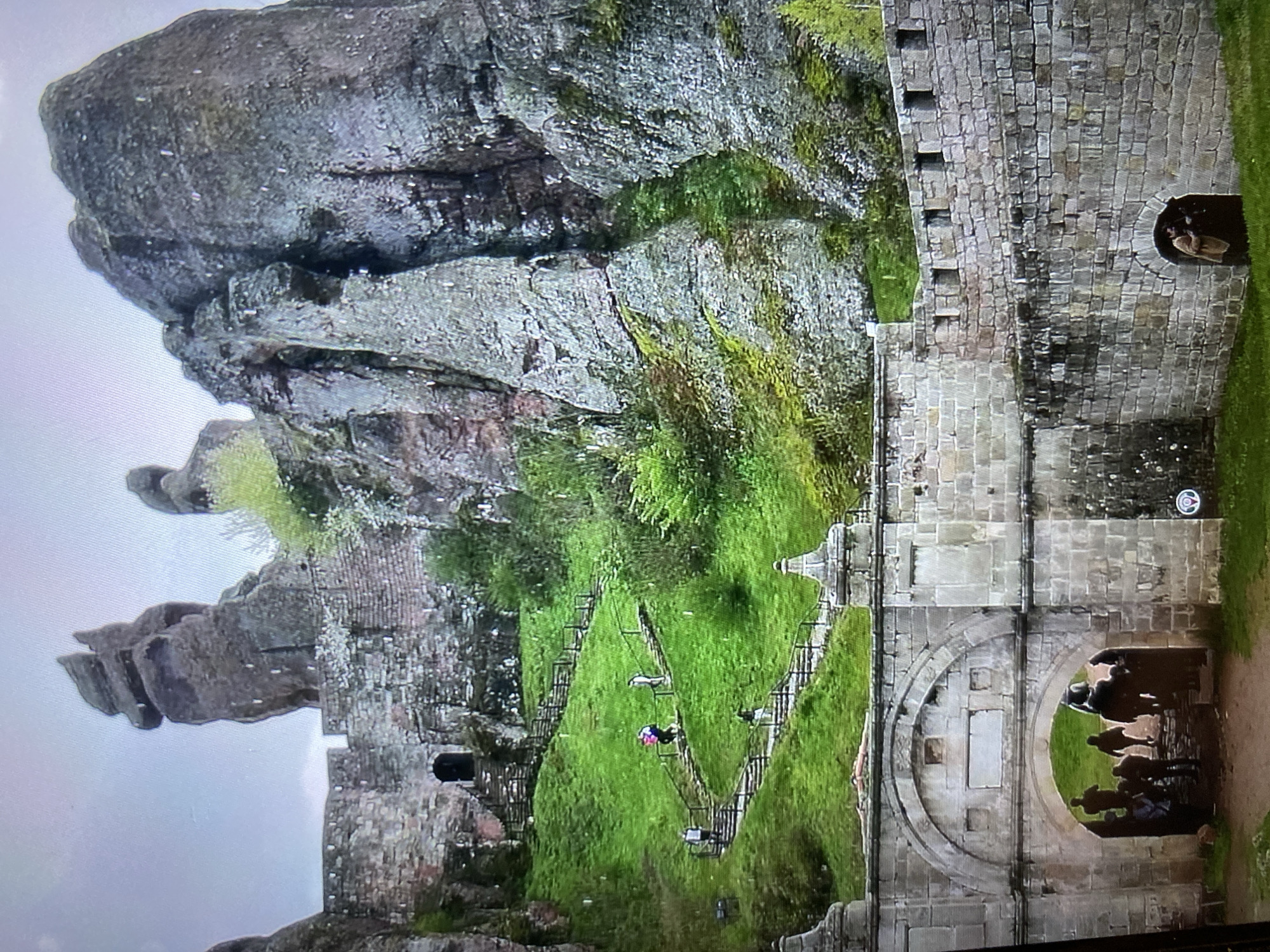 This image, correctly oriented, displays a striking outdoor scene dominated by natural rock formations and ancient-looking stone architecture, possibly a fortress or monastery.
On the left, towering grey rock cliffs rise dramatically, characterized by their rugged, weathered surfaces and horizontal striations. Patches of vibrant green moss and small vegetation cling to crevices and ledges on these rocks, indicating a damp or verdant environment.
To the right, an imposing stone wall, built from cut rectangular blocks, extends vertically. This wall features a battlemented top with evenly spaced crenellations. Near the top right, a small arched opening in the wall frames a person looking out.
In the mid-ground, a grassy slope descends between the rocks and the wall. Several people are visible here, some navigating a metal walkway or bridge structure equipped with railings. One individual on this walkway wears a distinctive pink backpack.
At the bottom right, the stone wall culminates in a large, grand arched gateway. Beneath this archway, several individuals are visible as dark silhouettes, either entering or exiting. The arch itself is ornately detailed with concentric stone layers and features a rectangular stone slab embedded above the arch, though no legible text is visible on it. A small, circular white sign with a red symbol is also affixed to the wall above the arch.
The lighting suggests an overcast or cloudy day, contributing to the diffused light and lack of strong shadows. The overall setting is a historical or natural heritage site, likely popular for exploration or tourism. The image doesn't contain specific textual cues that identify its location as Bankya, Bulgaria, but the combination of natural rock formations and ancient stone structures is consistent with such a region.